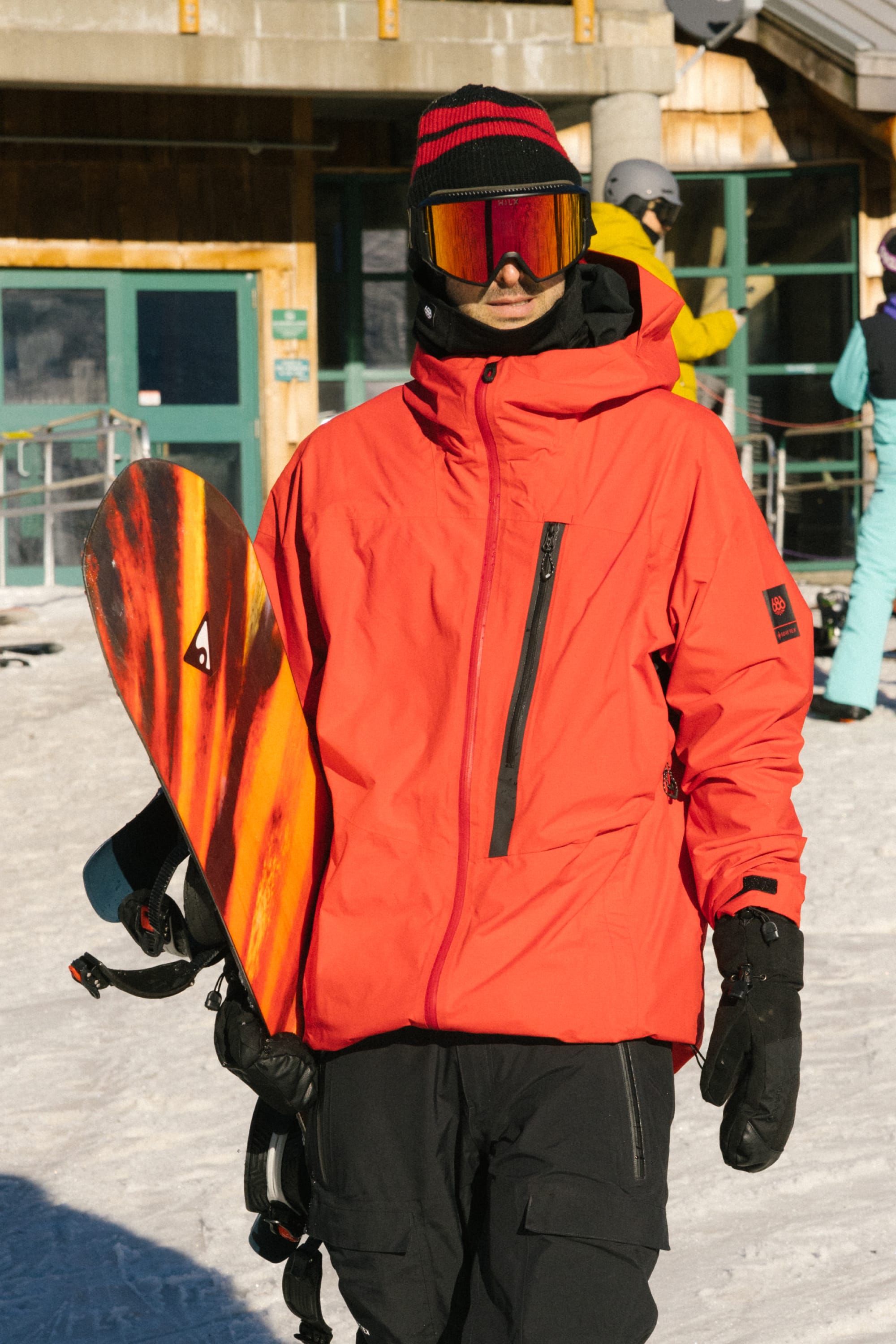 Wearing the 686 Men's GORE-TEX GT Thermagraph® Jacket, a person in black pants and mirrored goggles holds a red and orange snowboard while standing on snow near a ski lodge.