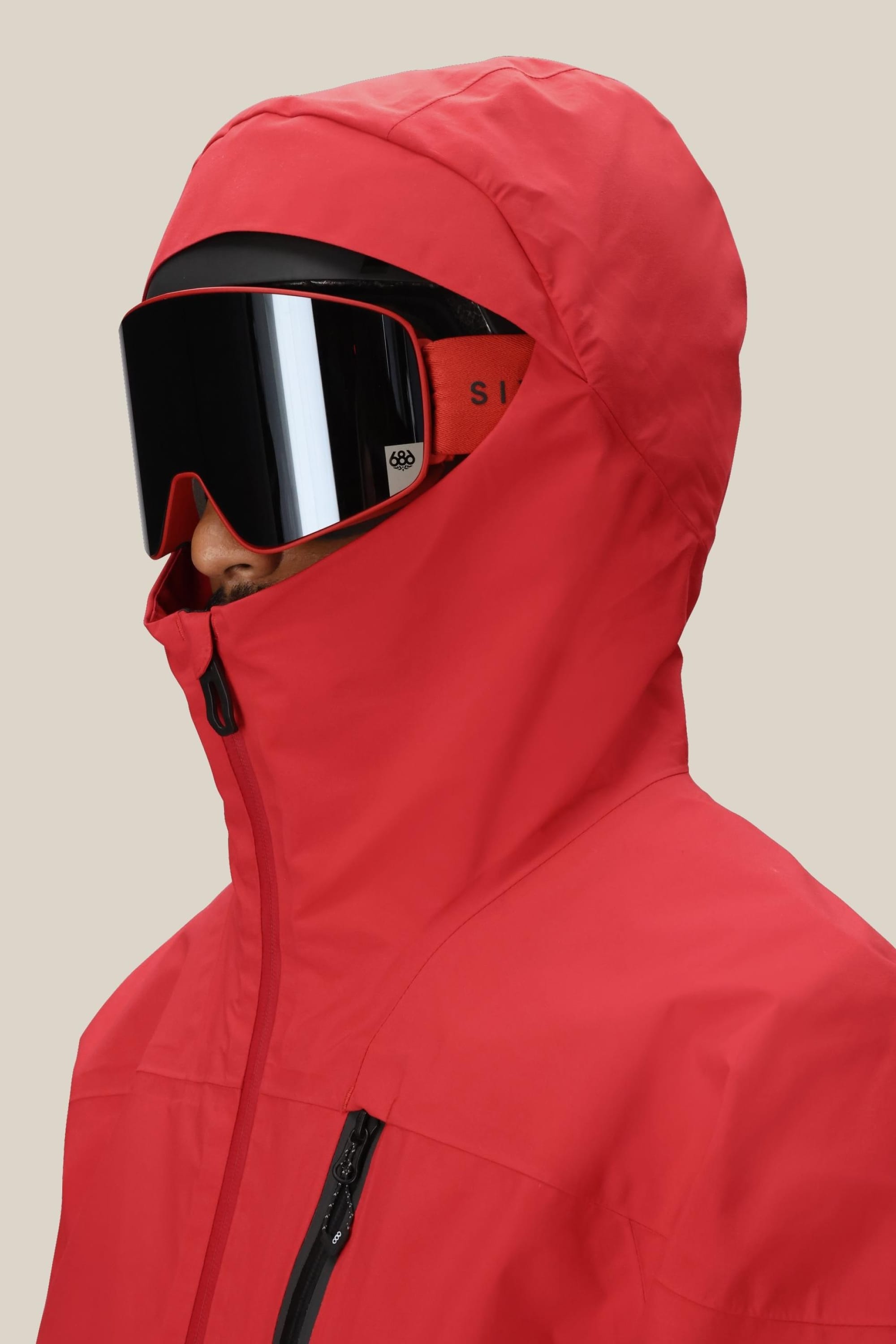 A person wears the 686 Men's GORE-TEX GT Thermagraph® Jacket in bright red, zipped up to the nose, paired with large black ski goggles. The background is plain and light-colored.