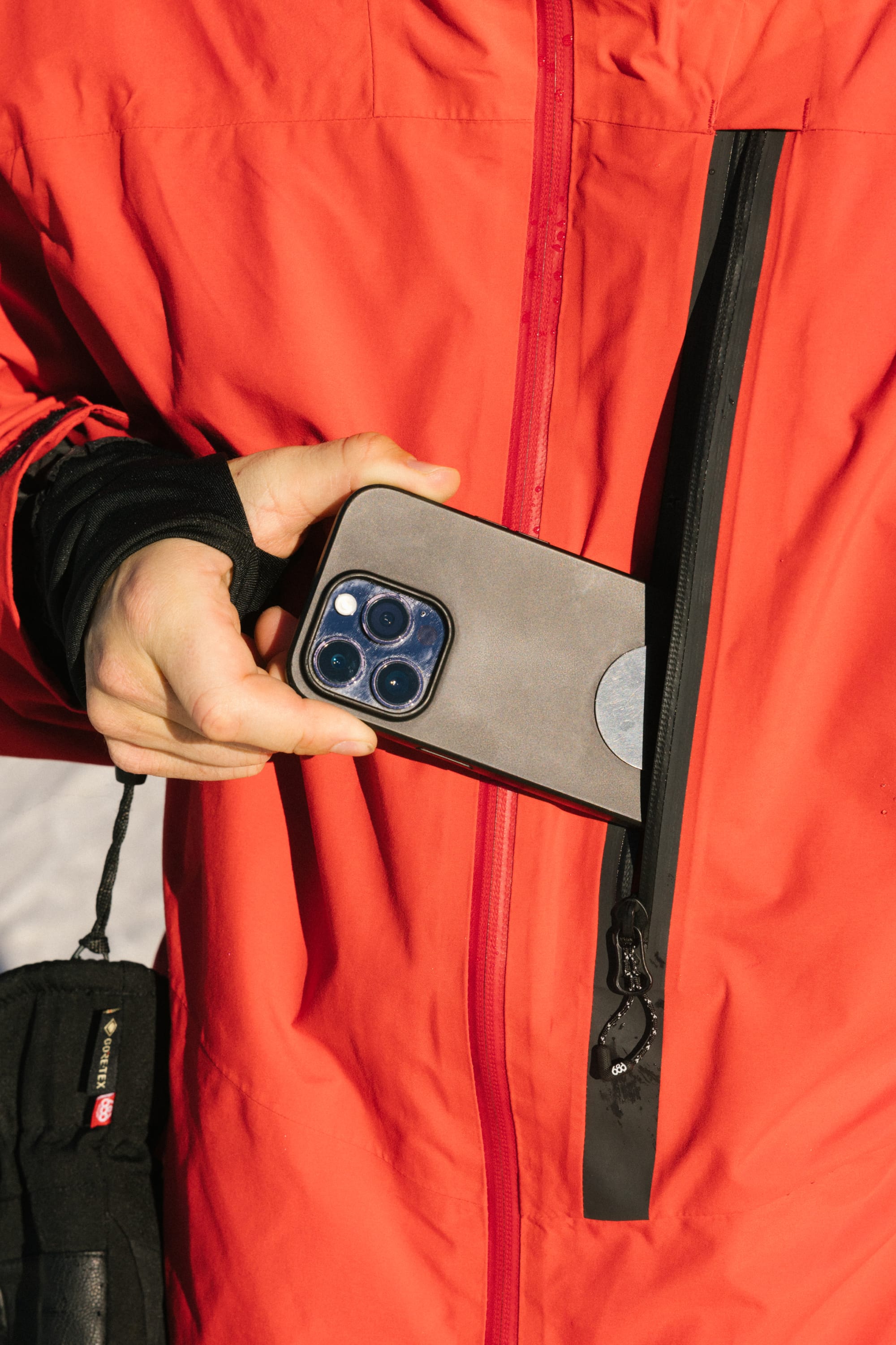 A man in a bright red 686 Men's GORE-TEX GT Thermagraph® Jacket unzips the vertical chest pocket and partially places a smartphone with a gray case inside.
