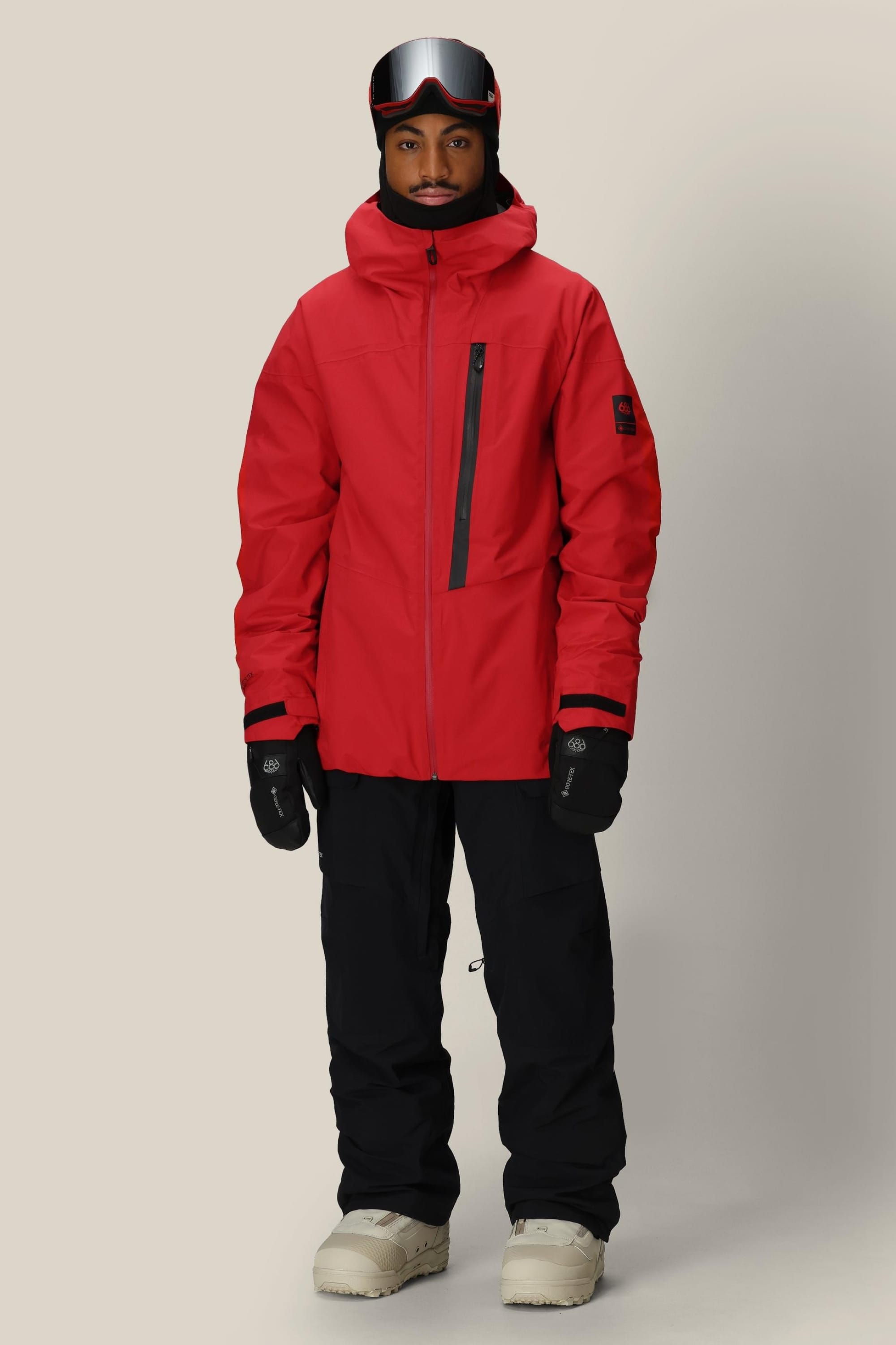 A man faces forward wearing the 686 Men's GORE-TEX GT Thermagraph® Jacket in red, with black snow pants, black gloves, white snow boots, and ski goggles. He has a neutral expression and stands against a plain background.