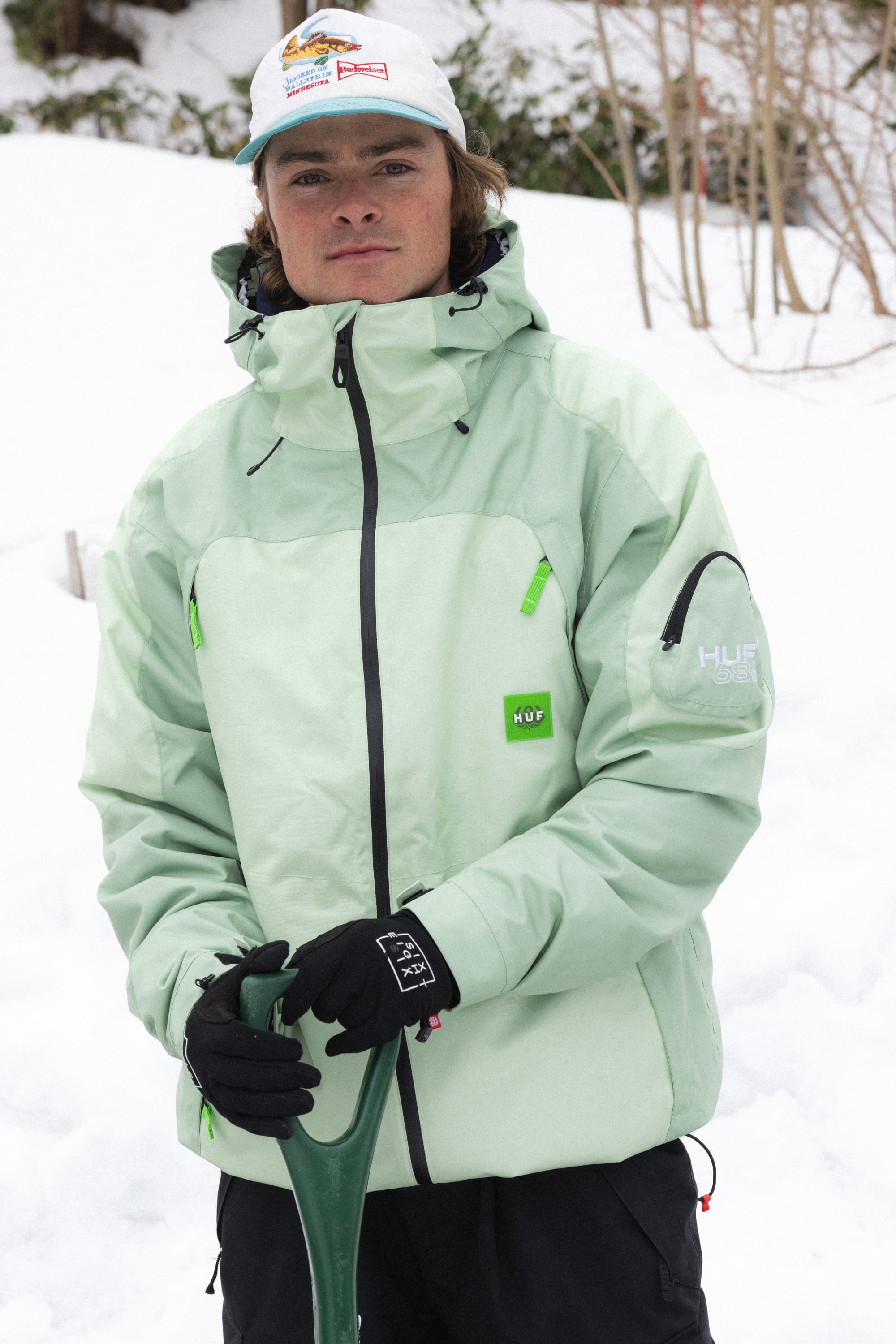 A person wearing the 686 Mens HUF Double H Shell Jacket, black gloves, and a white cap stands in the snow holding a green shovel, with bare trees in the background.