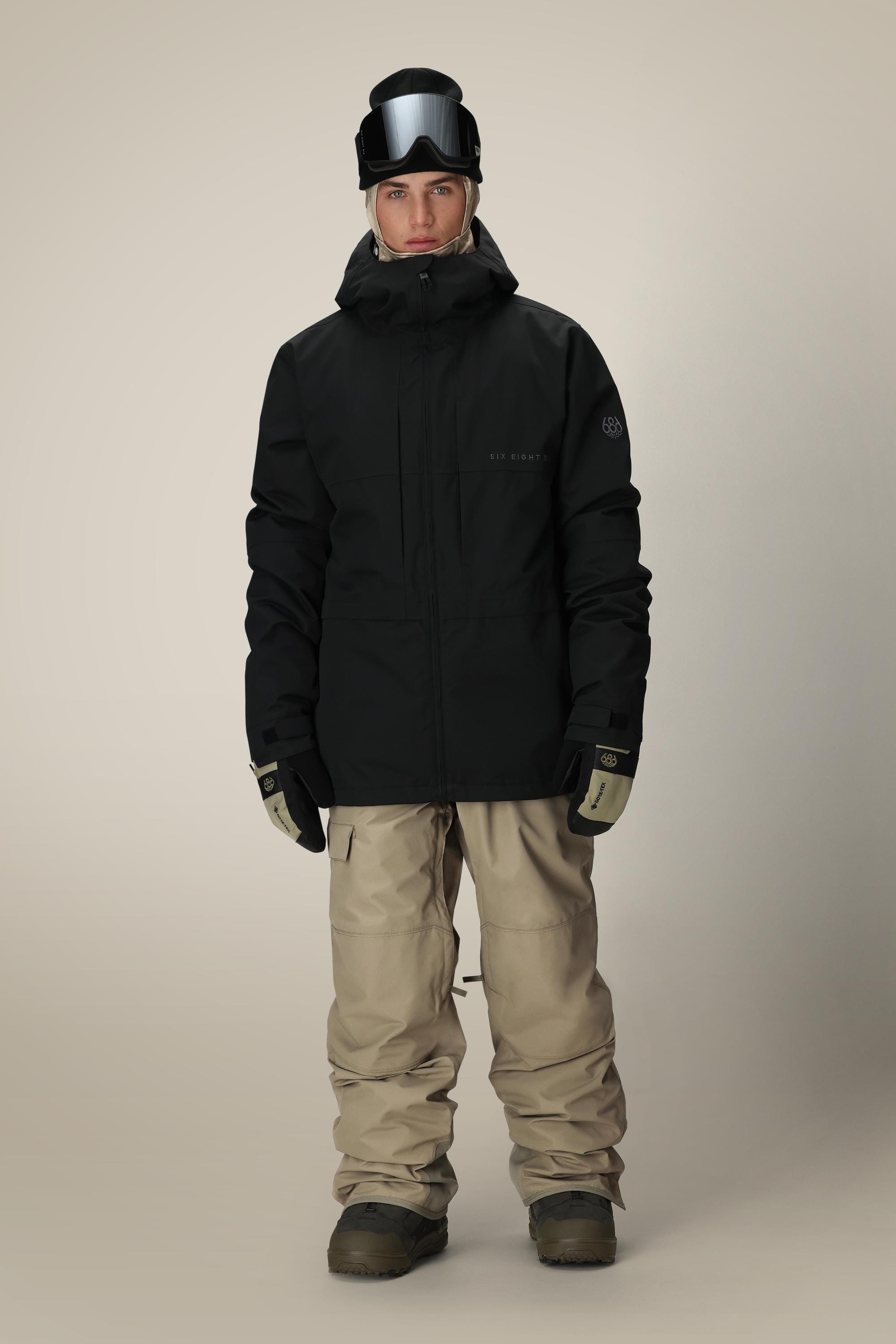A person wearing the 686 Men's SMARTY® 3-In-1 Form Jacket, beige snow pants, gloves, boots, and a black helmet with goggles stands facing forward against a plain light background.