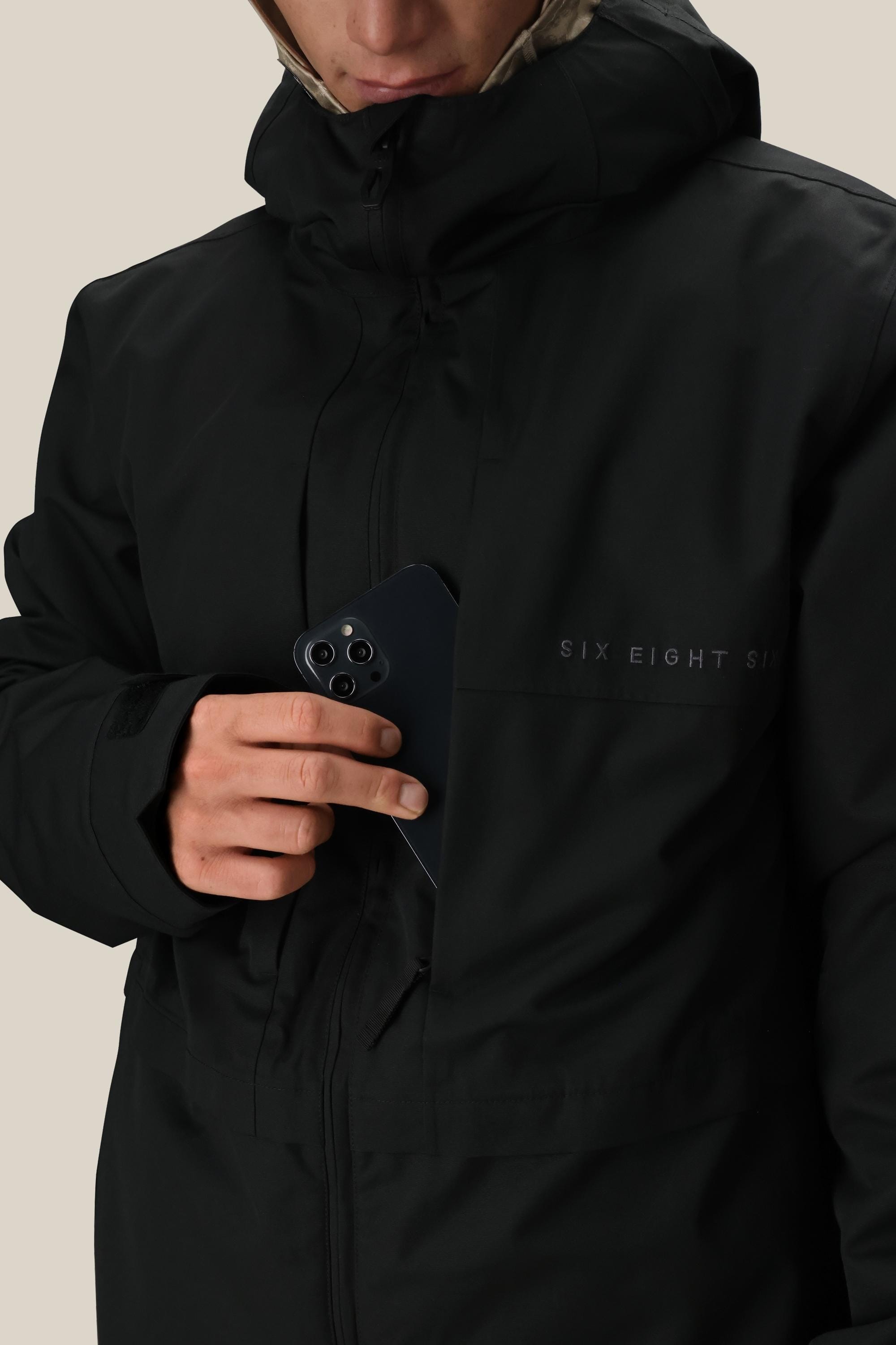 A person wearing the 686 Men’s SMARTY® 3-In-1 Form Jacket in black places a black smartphone into the chest pocket. Only the lower half of their face is visible.