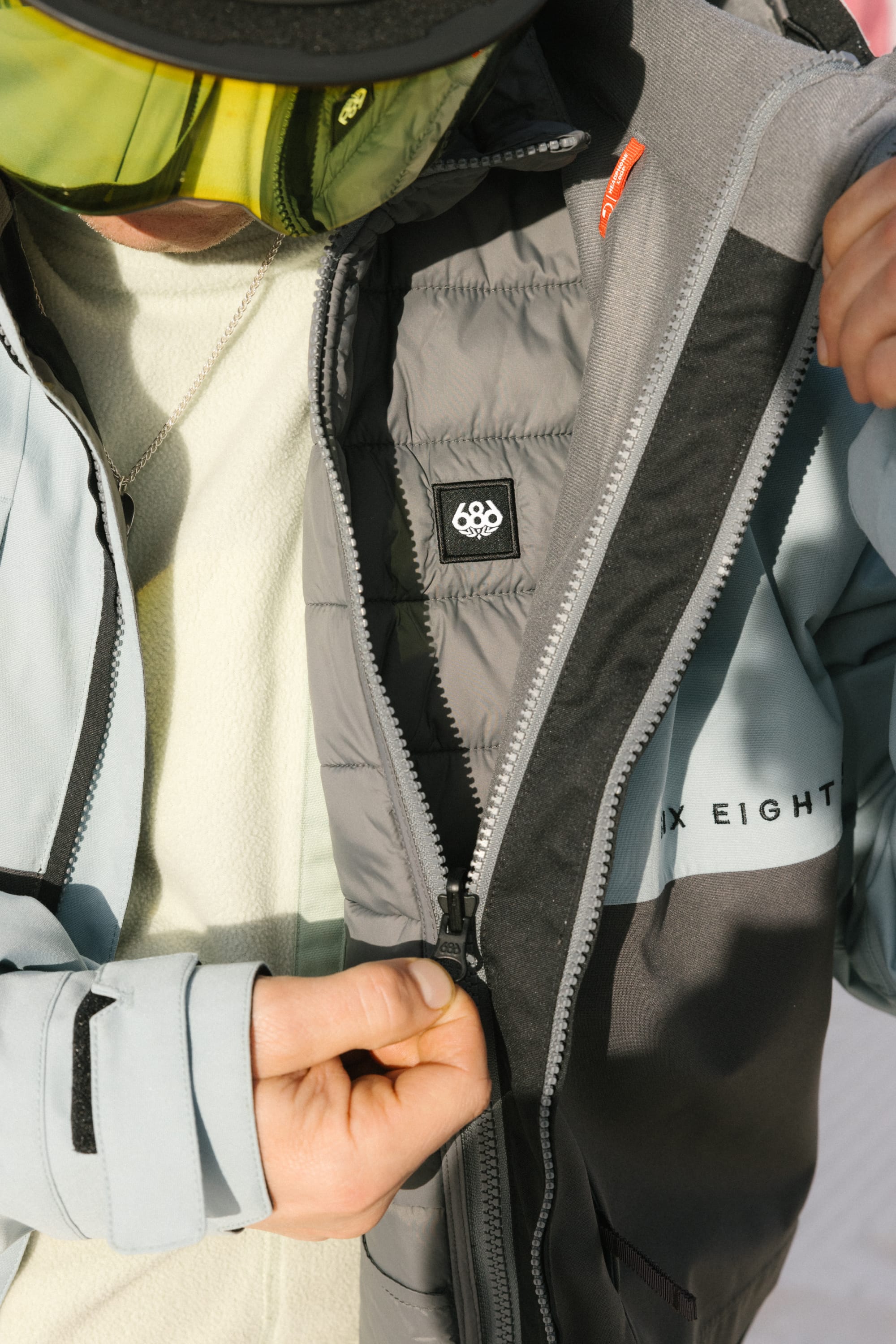 A man in ski goggles unzips his light blue-grey 686 Men's SMARTY® 3-In-1 Form Jacket, revealing the inner lining with the 686 logo and SIX EIGHT text on the chest.
