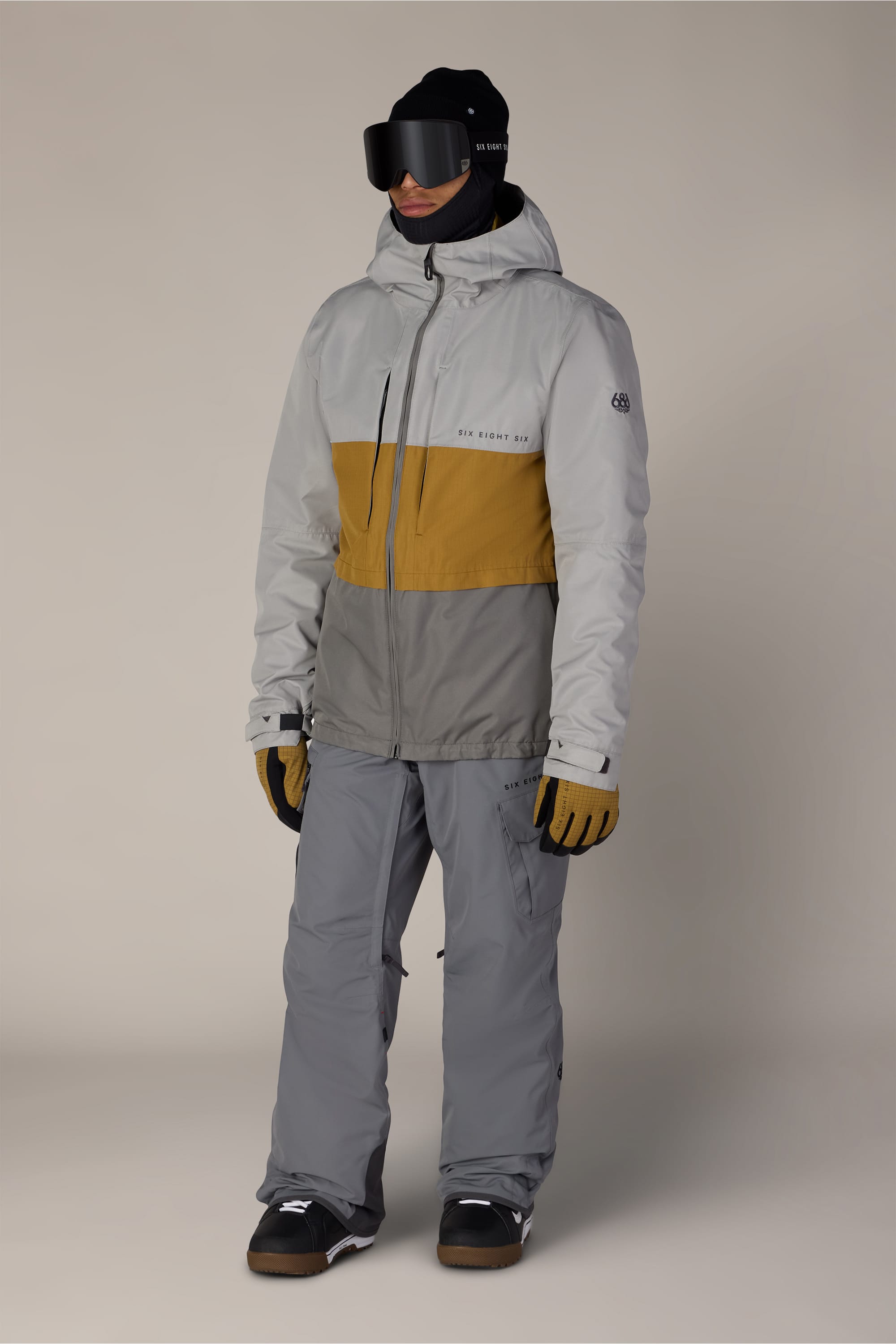 A person wearing the 686 Men’s SMARTY® 3-In-1 Form Jacket by 686 in gray and mustard yellow, along with gray snow pants, gloves, black boots, a black face covering, and black ski goggles stands against a plain background.