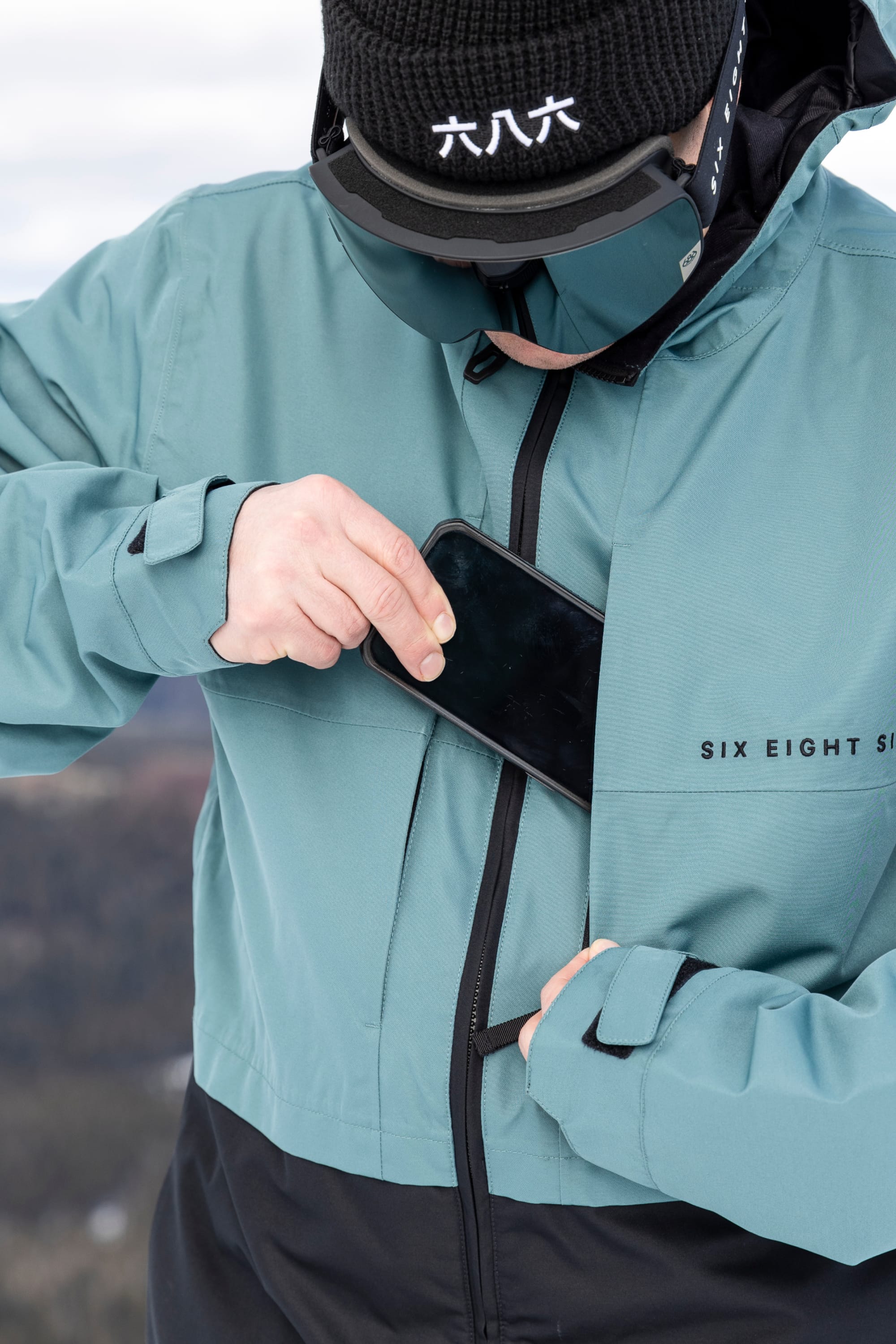 A person wearing the 686 Men’s SMARTY® 3-In-1 Form Jacket in teal and a black beanie slips a smartphone into the jacket’s chest pocket. Ski goggles rest on their forehead, while the outdoor background is softly blurred.