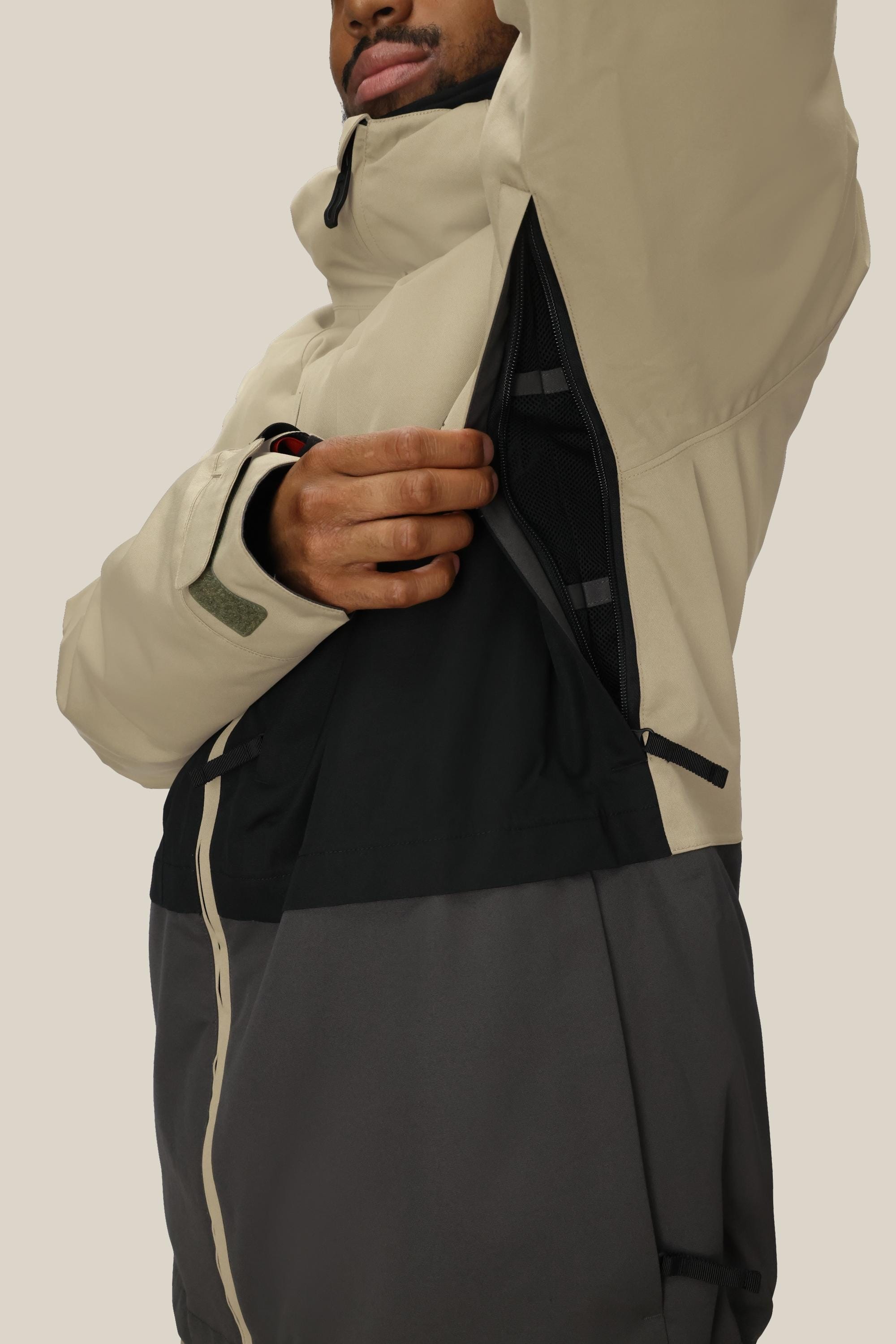 A person wearing the 686 Men's SMARTY® 3-In-1 Form Jacket in beige and black unzips an underarm vent, showing the breathable mesh lining. The two-tone jacket by 686 has a high collar, and the person's face is partially visible.