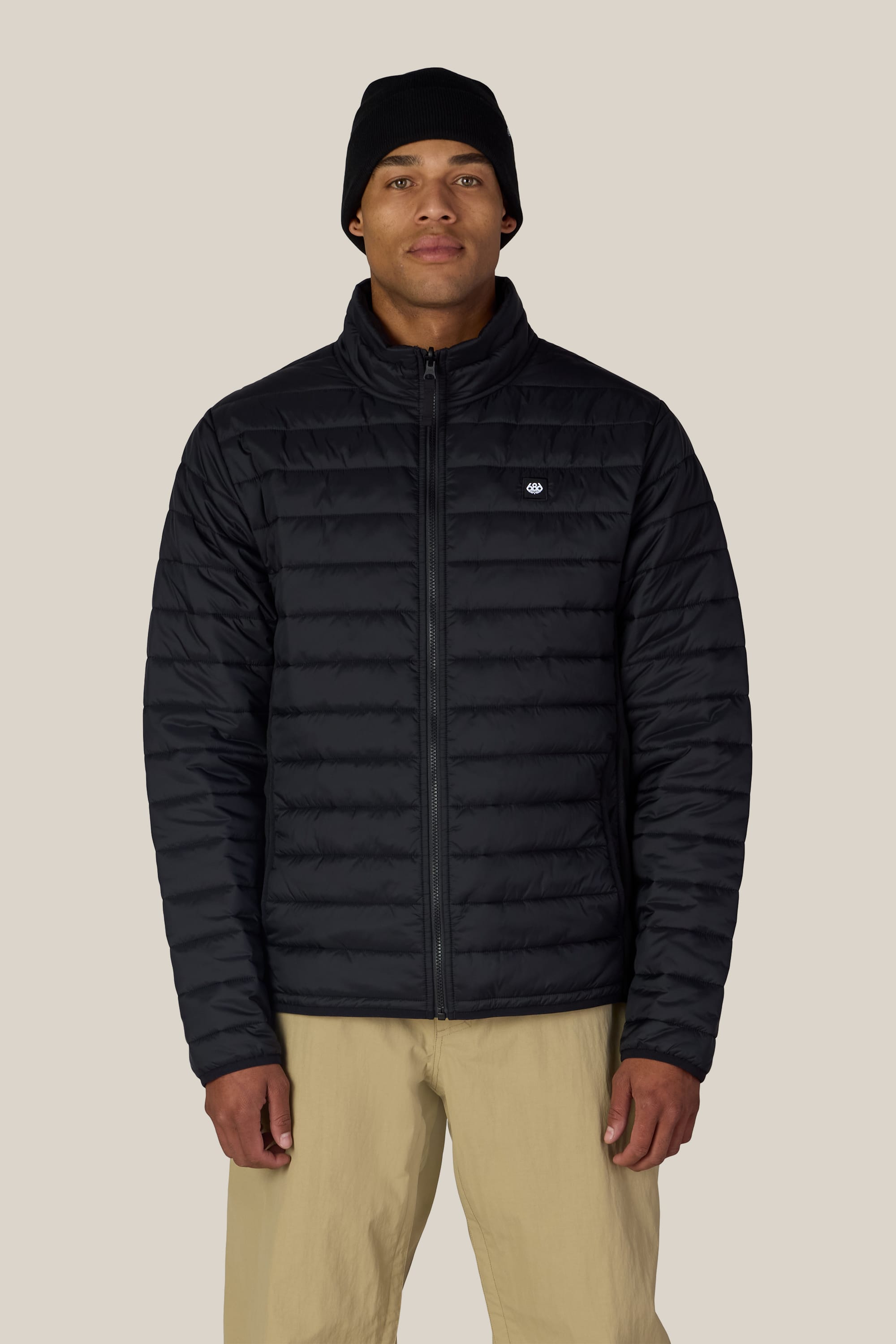 A person wearing a black beanie, beige pants, and the 686 Men’s SMARTY® 3-In-1 Form Jacket by 686—featuring a small logo on the left chest—stands facing the camera against a plain, light-colored background.