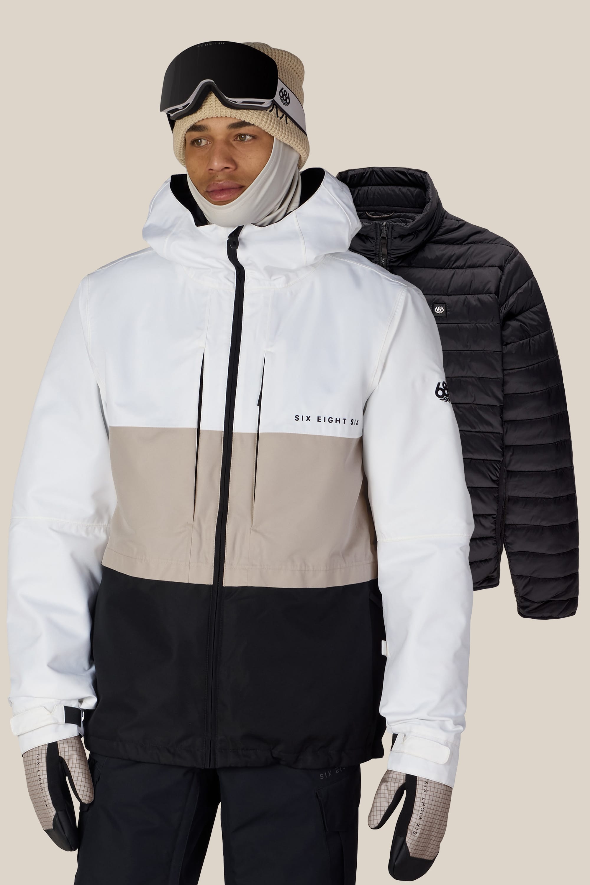 A person wearing the 686 Men’s SMARTY® 3-In-1 Form Jacket by 686, along with gloves, a beige knit hat, and ski goggles, stands in front of a black puffer jacket on a neutral background.