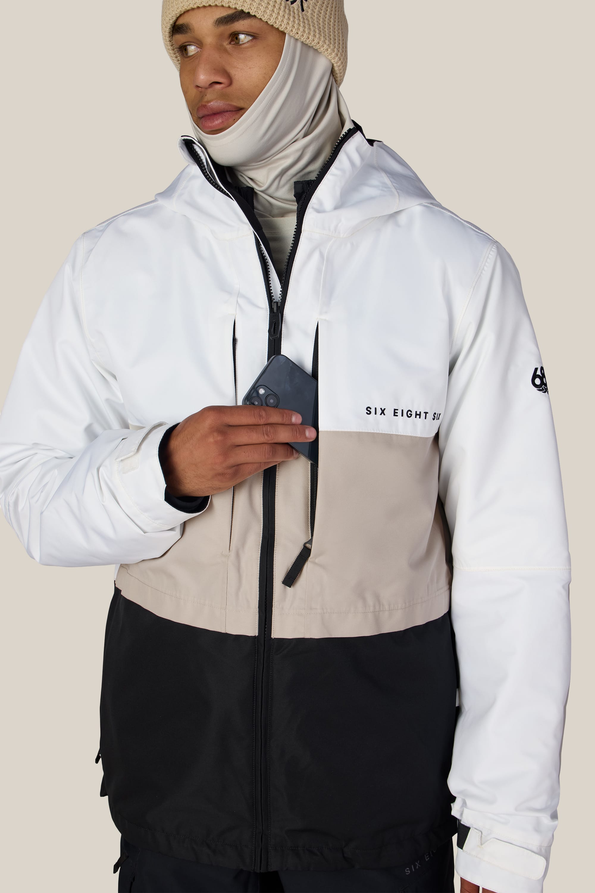 A man wearing the 686 Men’s SMARTY® 3-In-1 Form Jacket in a white, beige, and black color-blocked design with “SIX EIGHT” text on the chest and a beige knit hat puts a card into his jacket pocket.