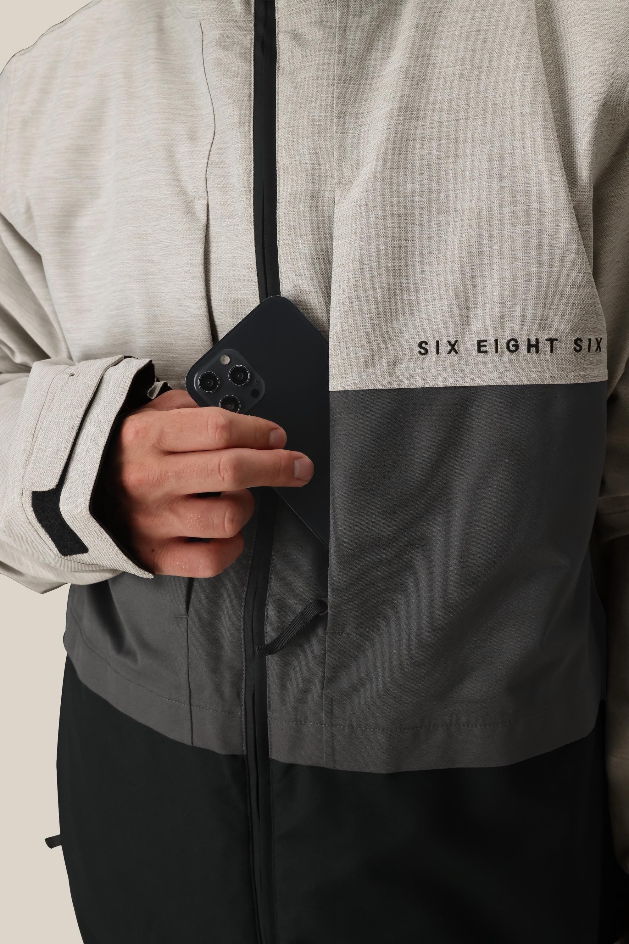 A person wearing the 686 Men's SMARTY® 3-In-1 Form Jacket in grey and black puts a black smartphone into the chest pocket.