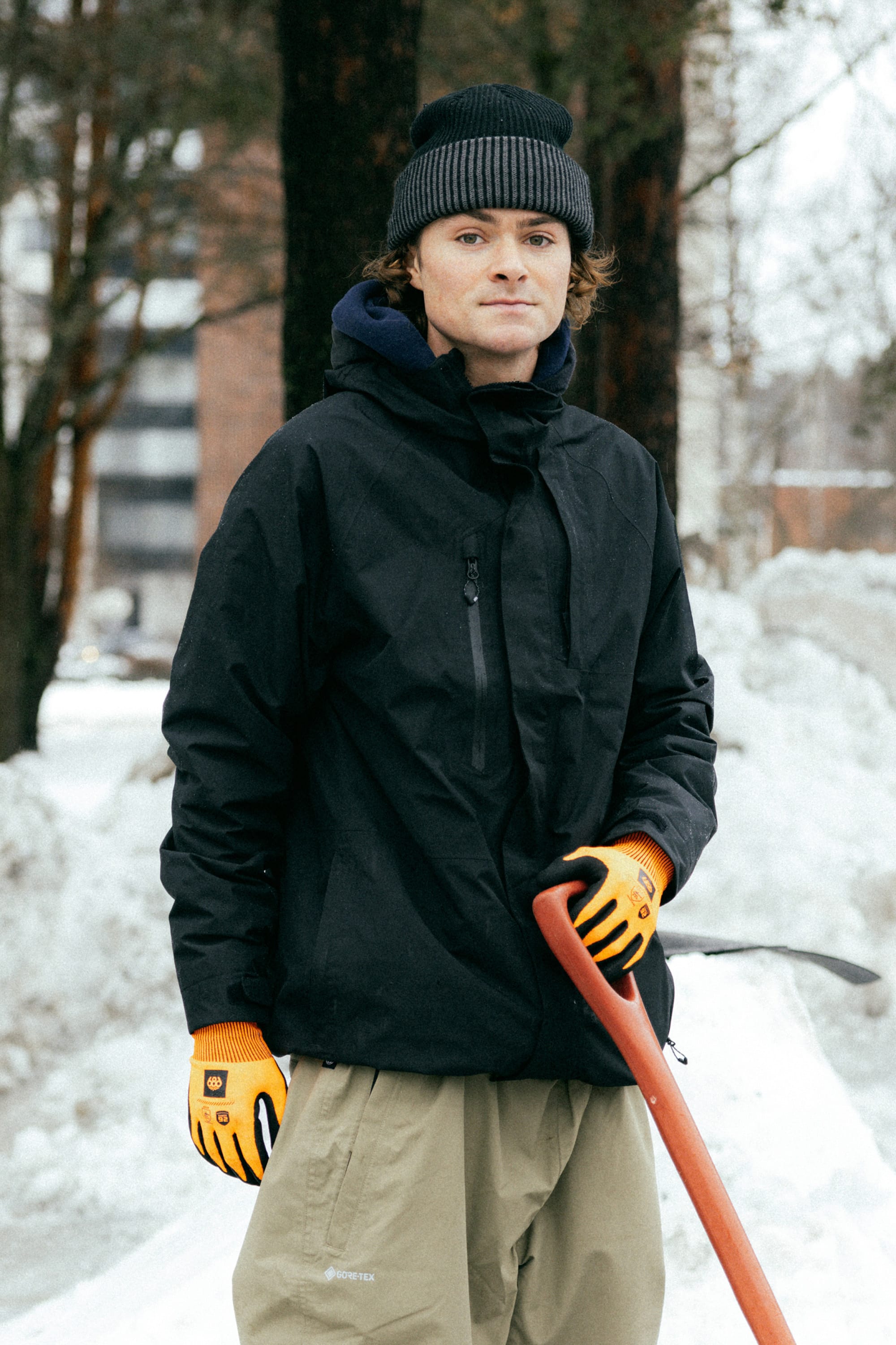 A person wearing the 686 Mens GORE-TEX Core Shell Jacket in black, beige pants, orange gloves, and a black knit beanie stands outside in the snow with a red-handled shovel; snow piles and trees are visible in the background.