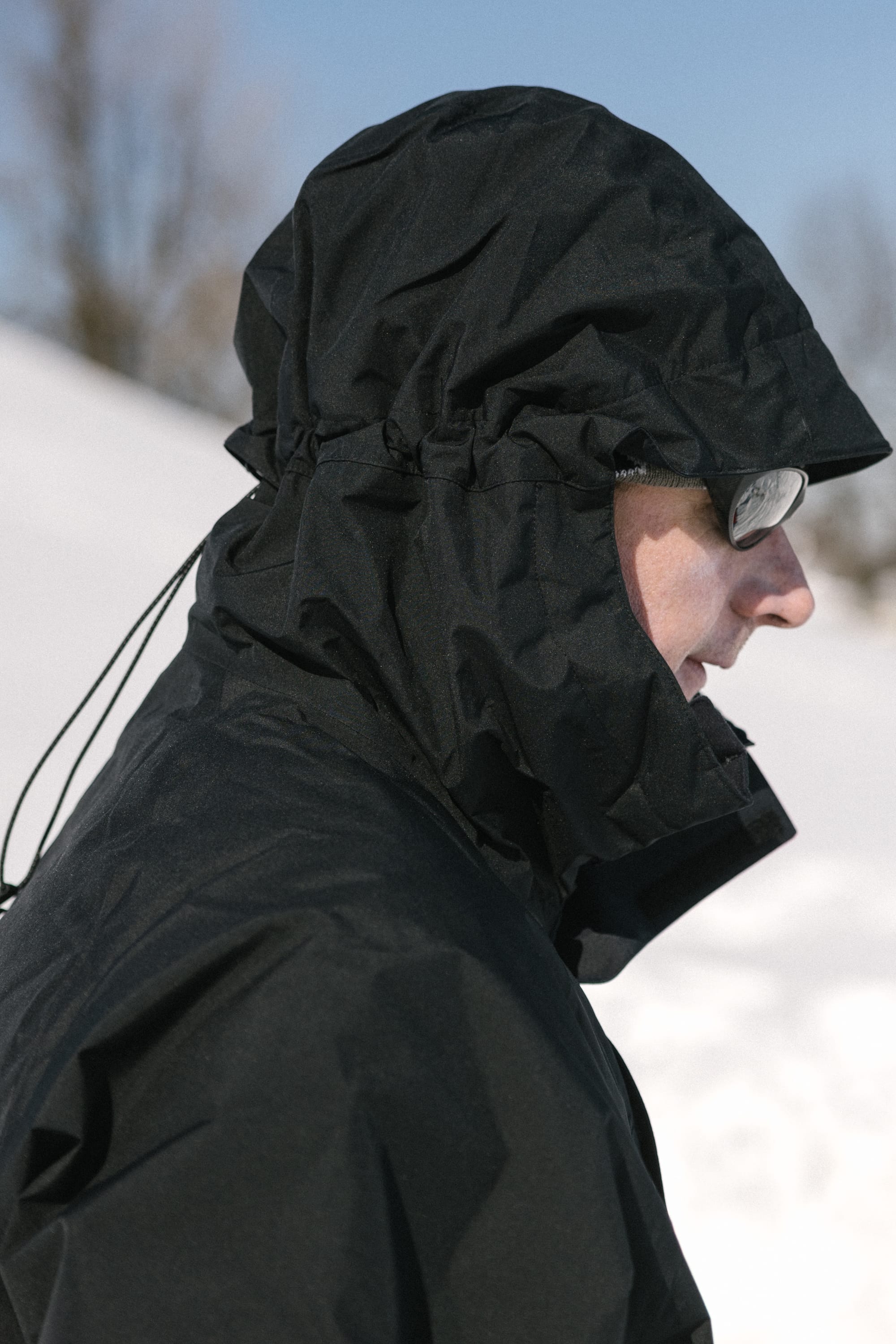 A person wearing the 686 Men’s GORE-TEX® Core Shell Jacket and glasses stands outdoors in a snowy landscape, with trees and a blue sky in the background.