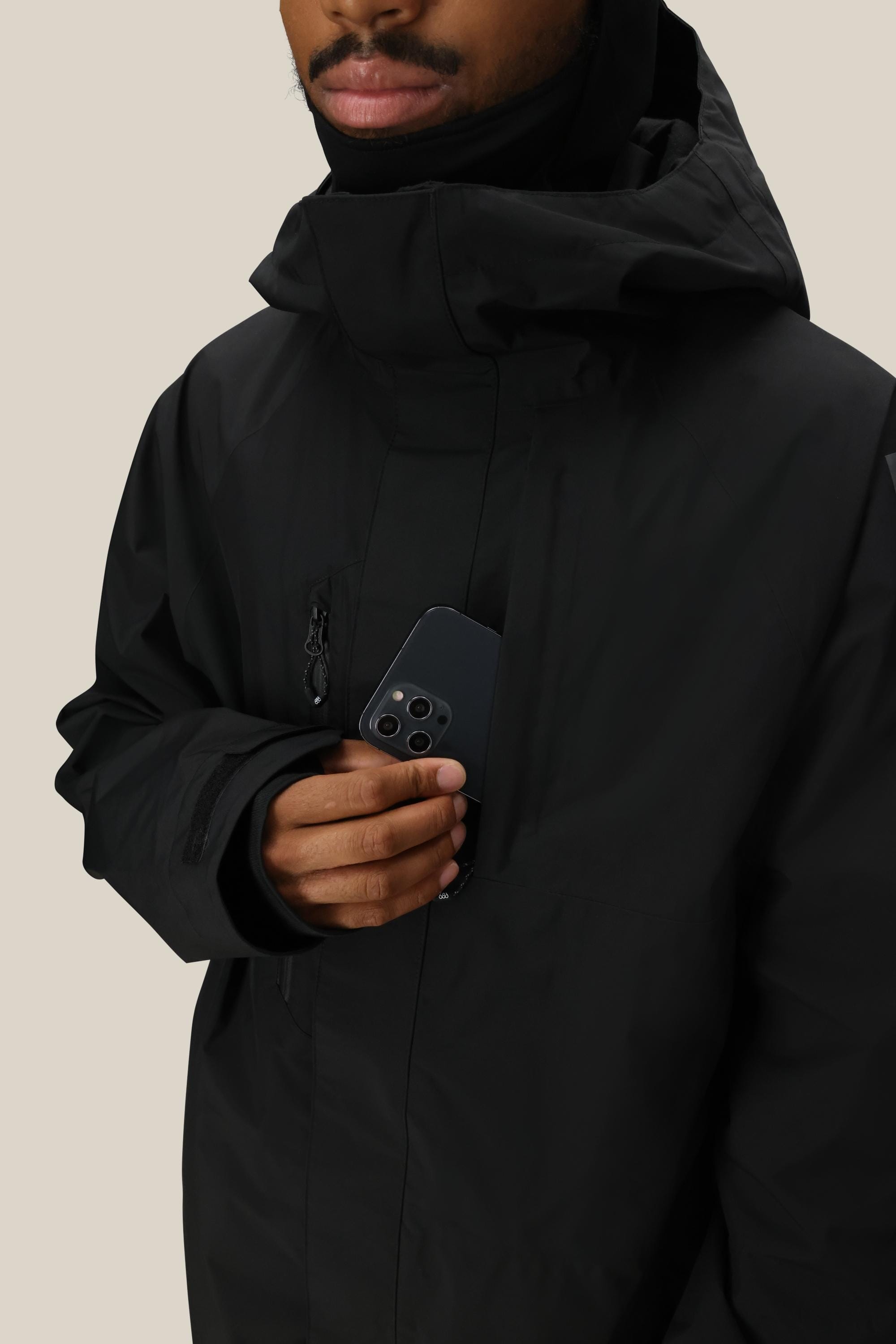 A man wearing a 686 Mens GORE-TEX Core Shell Jacket in black slips a black smartphone into its chest pocket. The background is plain beige, and only the lower half of his face is shown.