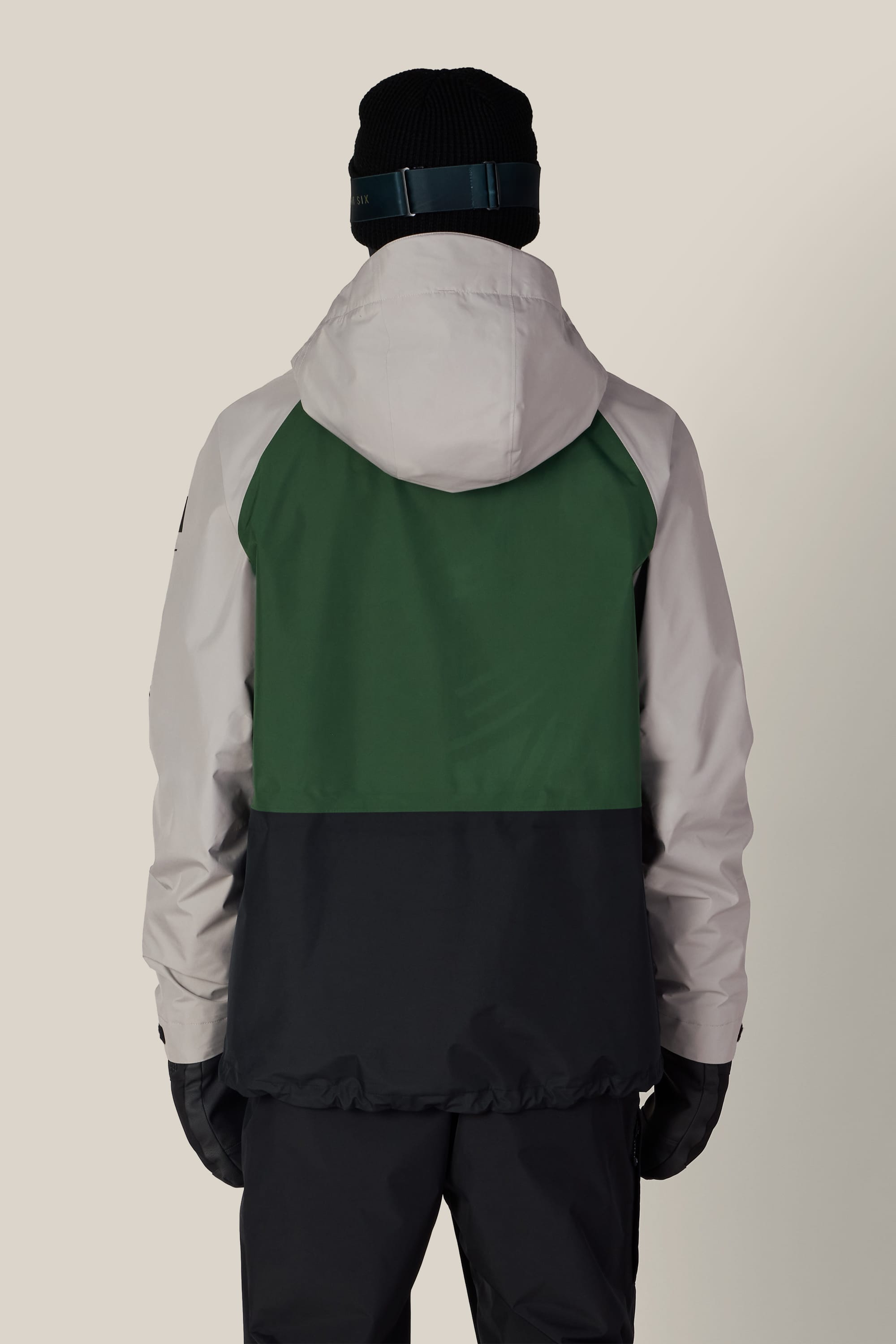 A person stands with their back to the camera against a plain background, wearing the 686 Men’s GORE-TEX® Core Shell Jacket by 686. The jacket features a color-blocked design with a white hood and sleeves, green upper back, and black lower back. They also wear a black beanie, dark pants, and gloves.