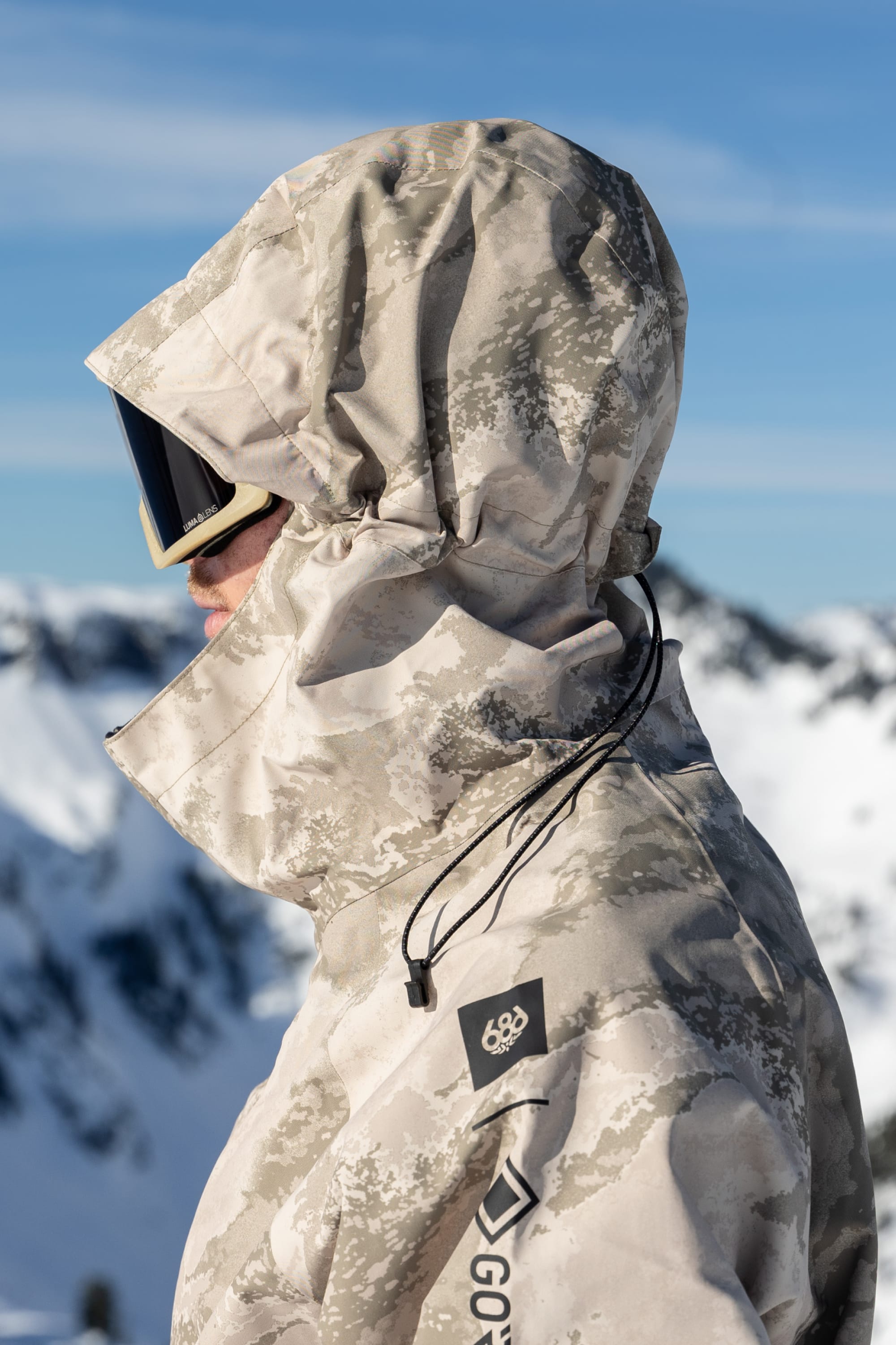 A man outdoor in snowy mountains, wearing the 686 Mens GORE-TEX Core Shell Jacket in a light camo pattern with hood and goggles, faces sideways against a blue sky.