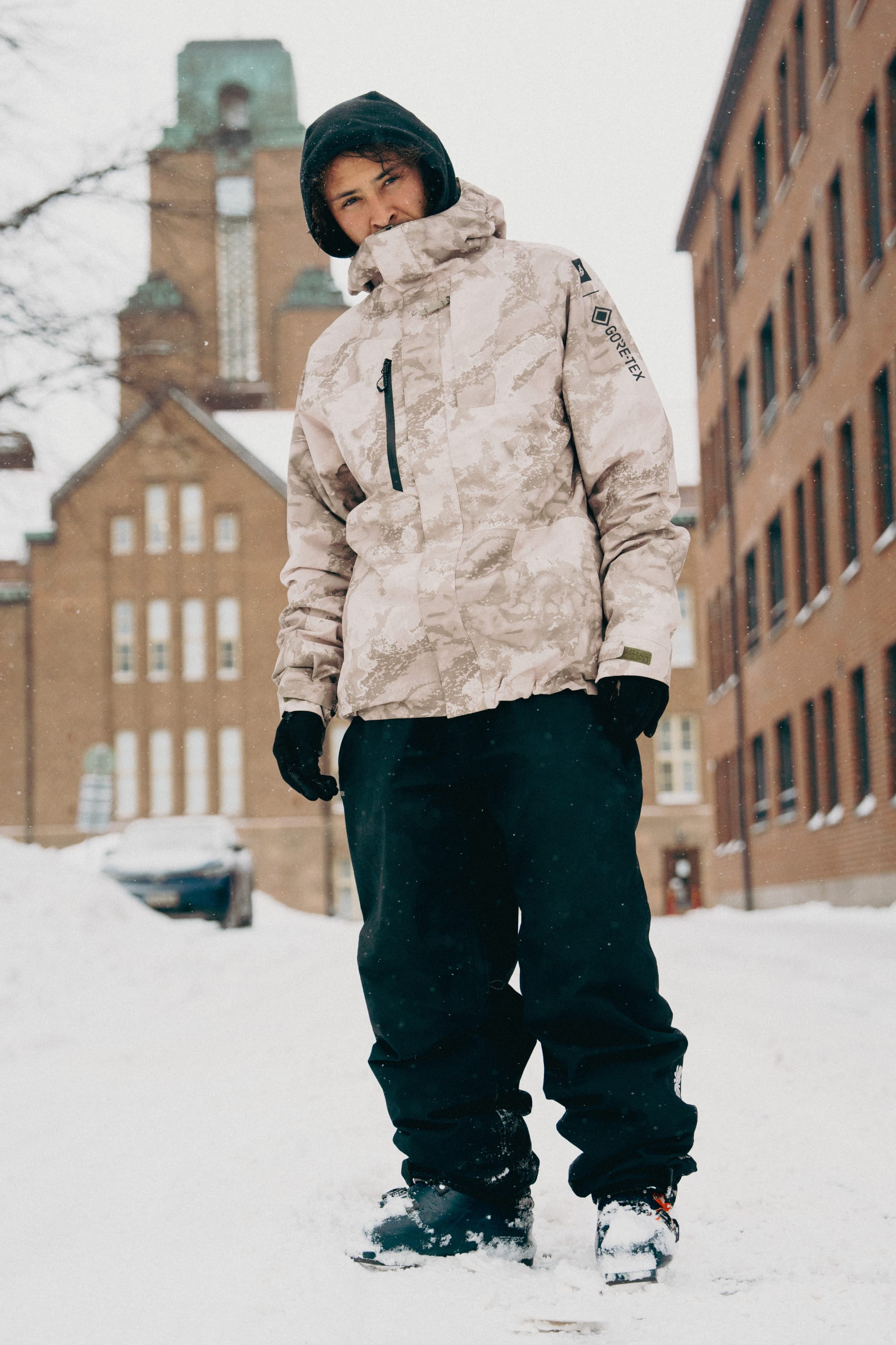 Wearing a 686 Mens GORE-TEX Core Shell Jacket with light camo, black snow pants, gloves, and a hood, a person stands on a snowy street between brick buildings as snow falls. A tall building with a tower is visible in the background.