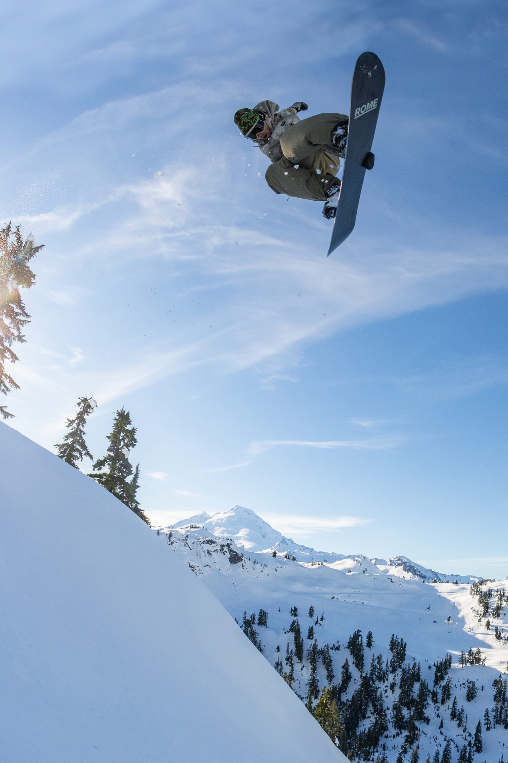 A snowboarder in a 686 Mens GORE-TEX Core Shell Jacket soars above a snowy slope dotted with pine trees and mountains, framed by a clear blue sky.