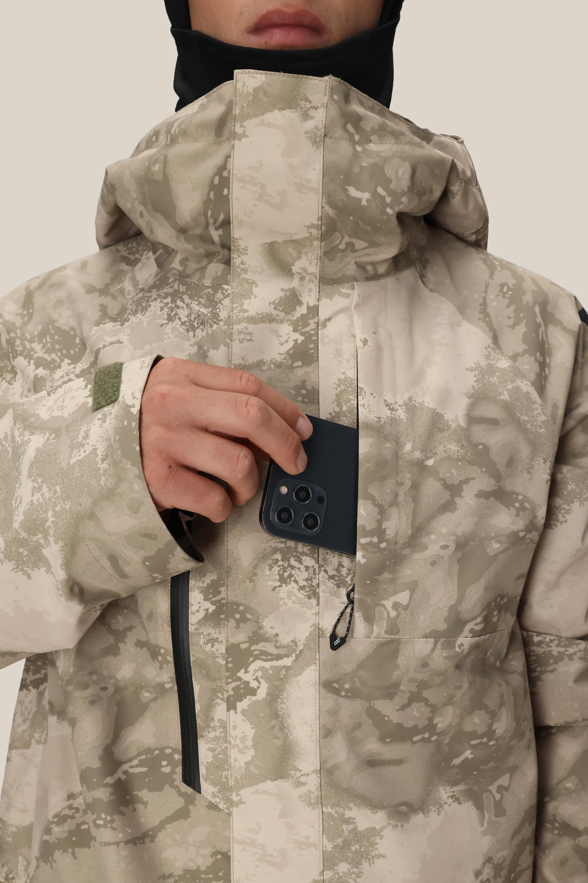 A person wearing the 686 Mens GORE-TEX Core Shell Jacket in beige with camouflage inserts places a black smartphone into the zippered chest pocket. The high-collar jacket by 686 blends style and function, with the persons face partially visible.