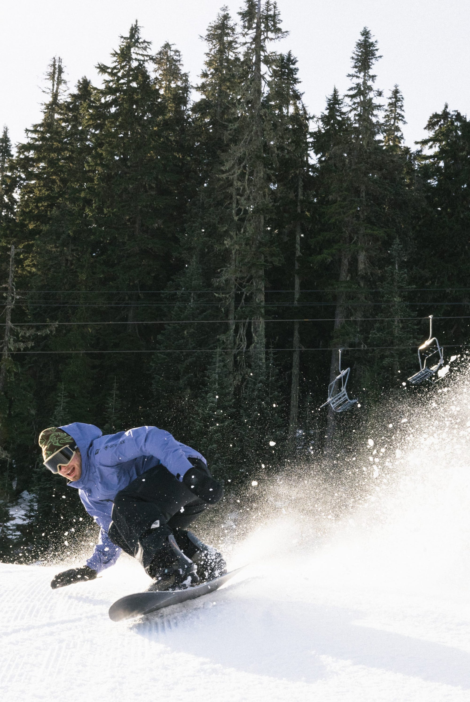 A snowboarder in a 686 Mens GORE-TEX Core Shell Jacket and black pants carves down a snowy slope, kicking up powder with tall evergreens and empty ski lift chairs in the background.