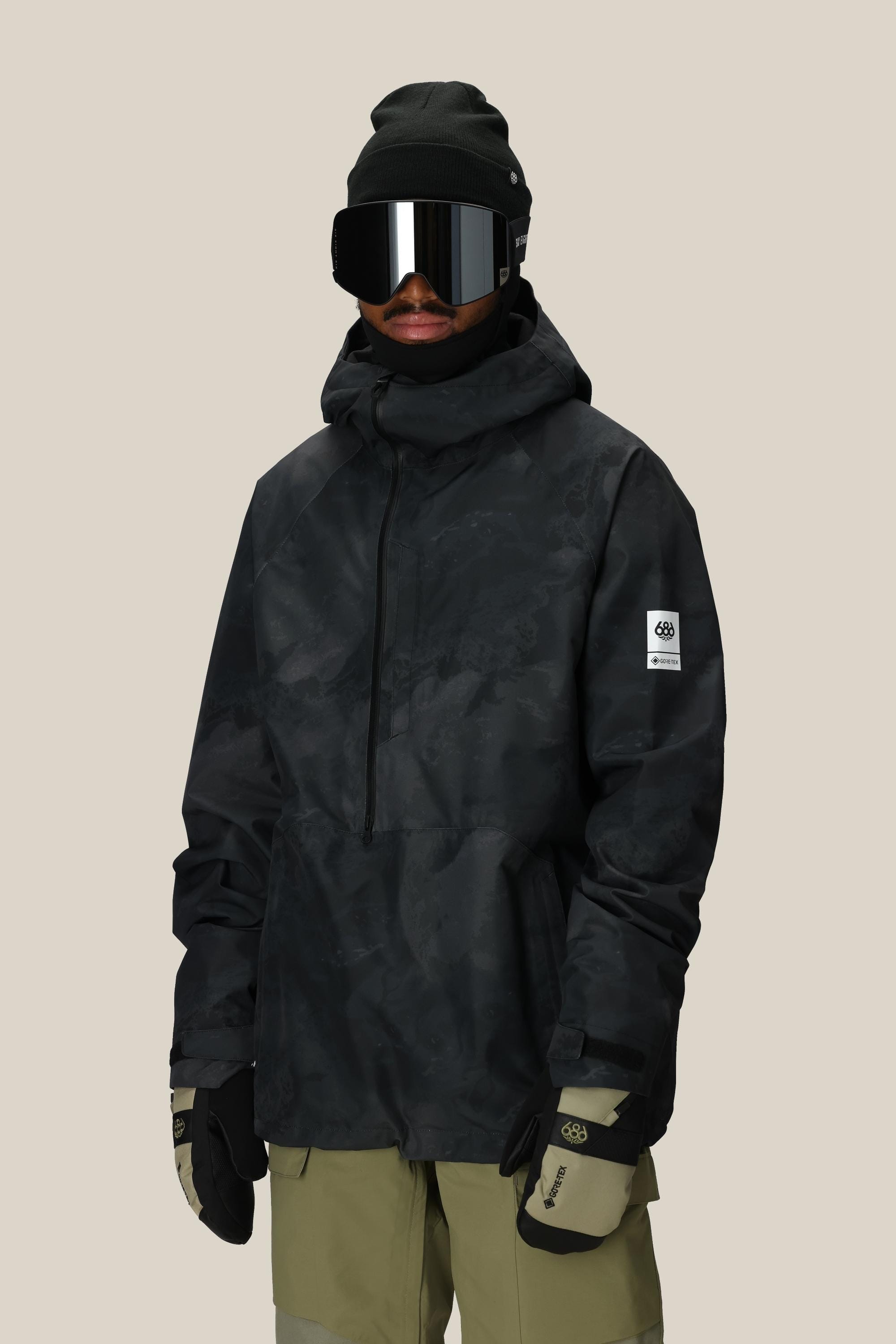 A person wearing a black beanie, large reflective ski goggles, beige gloves, olive green pants, and the 686 Mens GORE-TEX Fragment Shell Anorak stands against a plain light background.