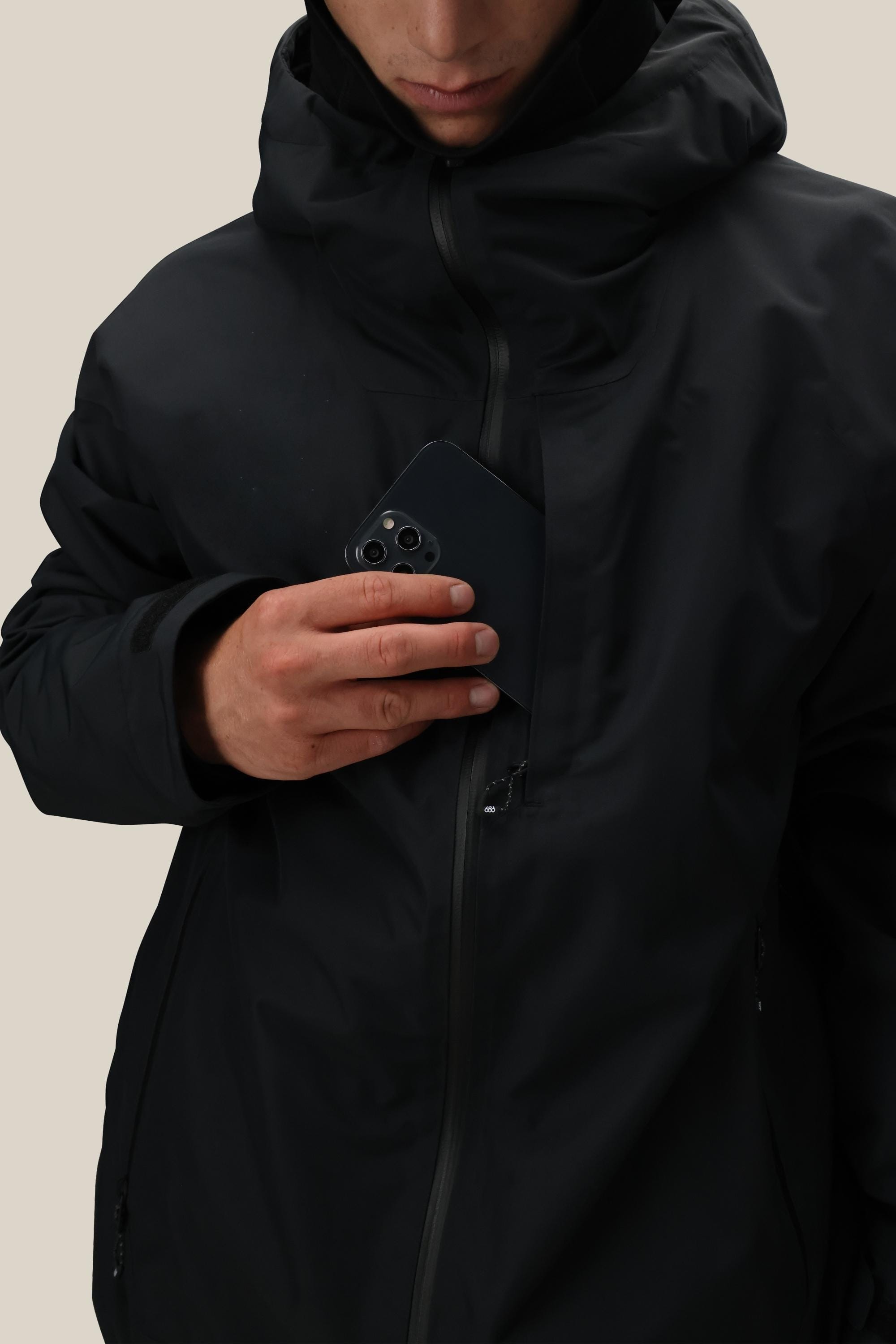 A man wearing the 686 Mens Gateway Shell Jacket in black places a black smartphone into its chest pocket, set against a plain light-colored background.