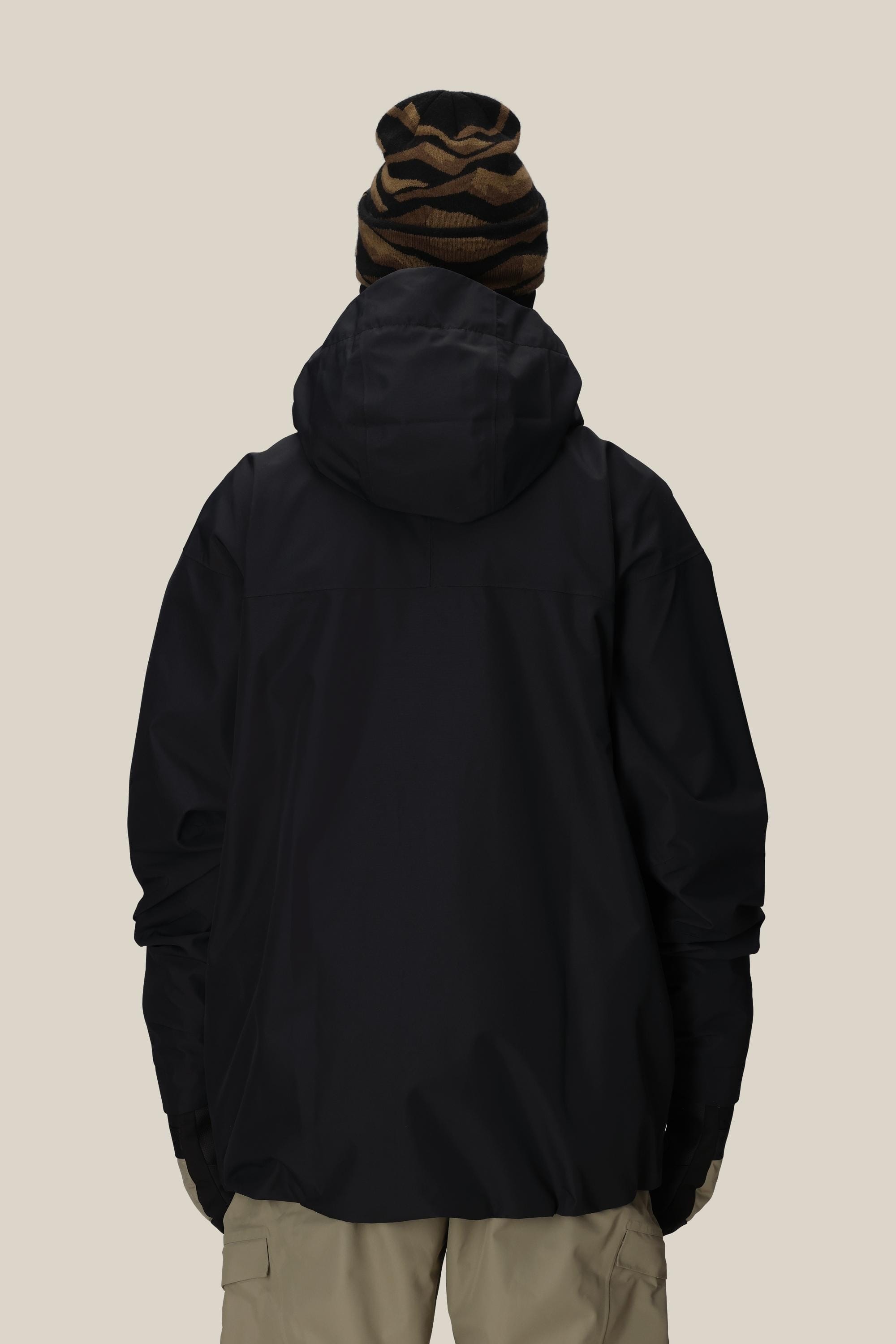 A person is shown from the back against a plain, light background wearing a 686 Mens Gateway Shell Jacket in black, beige pants, and a brown and black striped beanie.