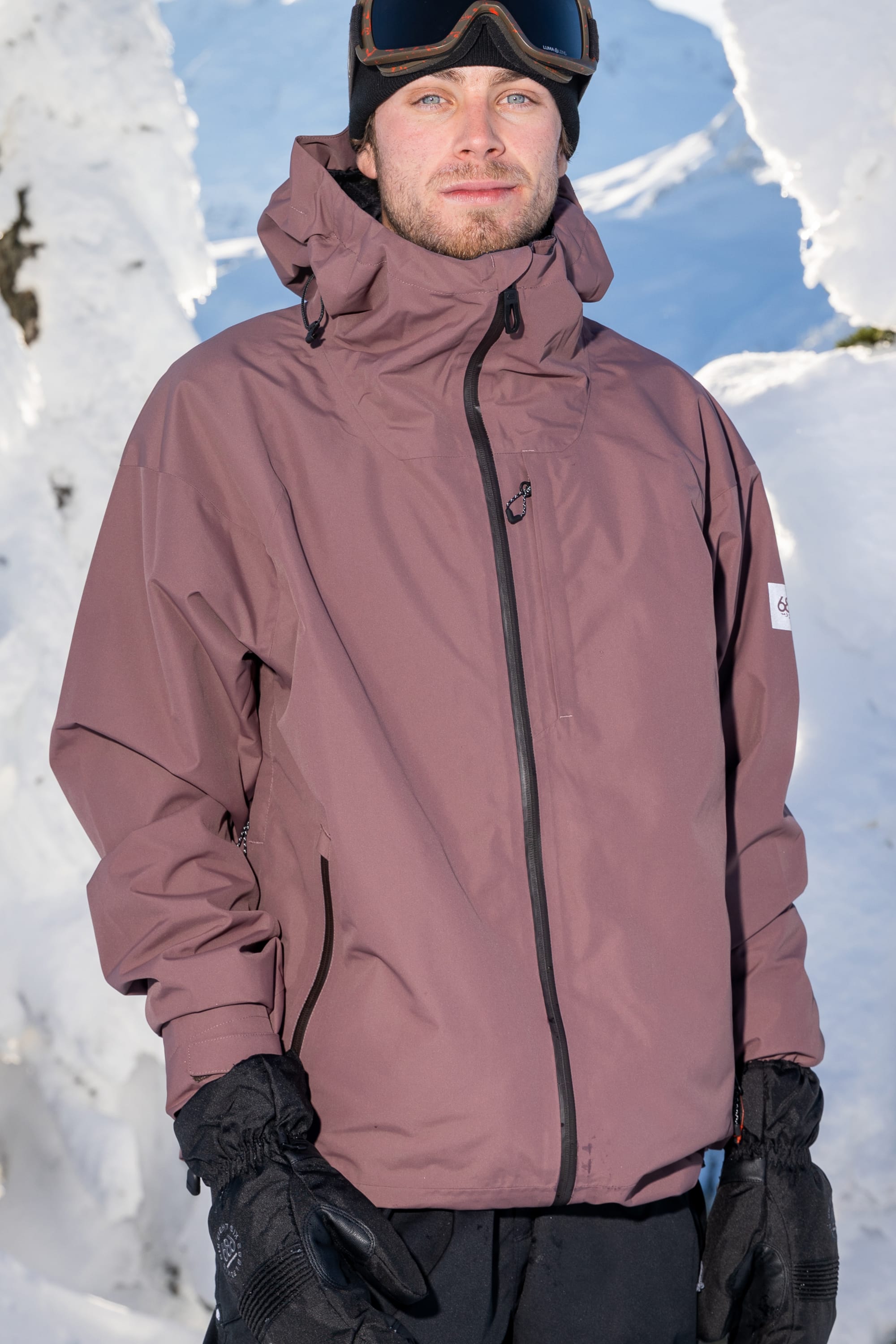 A person wearing the 686 Mens Gateway Shell Jacket by 686, along with black gloves and ski goggles, stands outdoors surrounded by snow and bright sunlight.