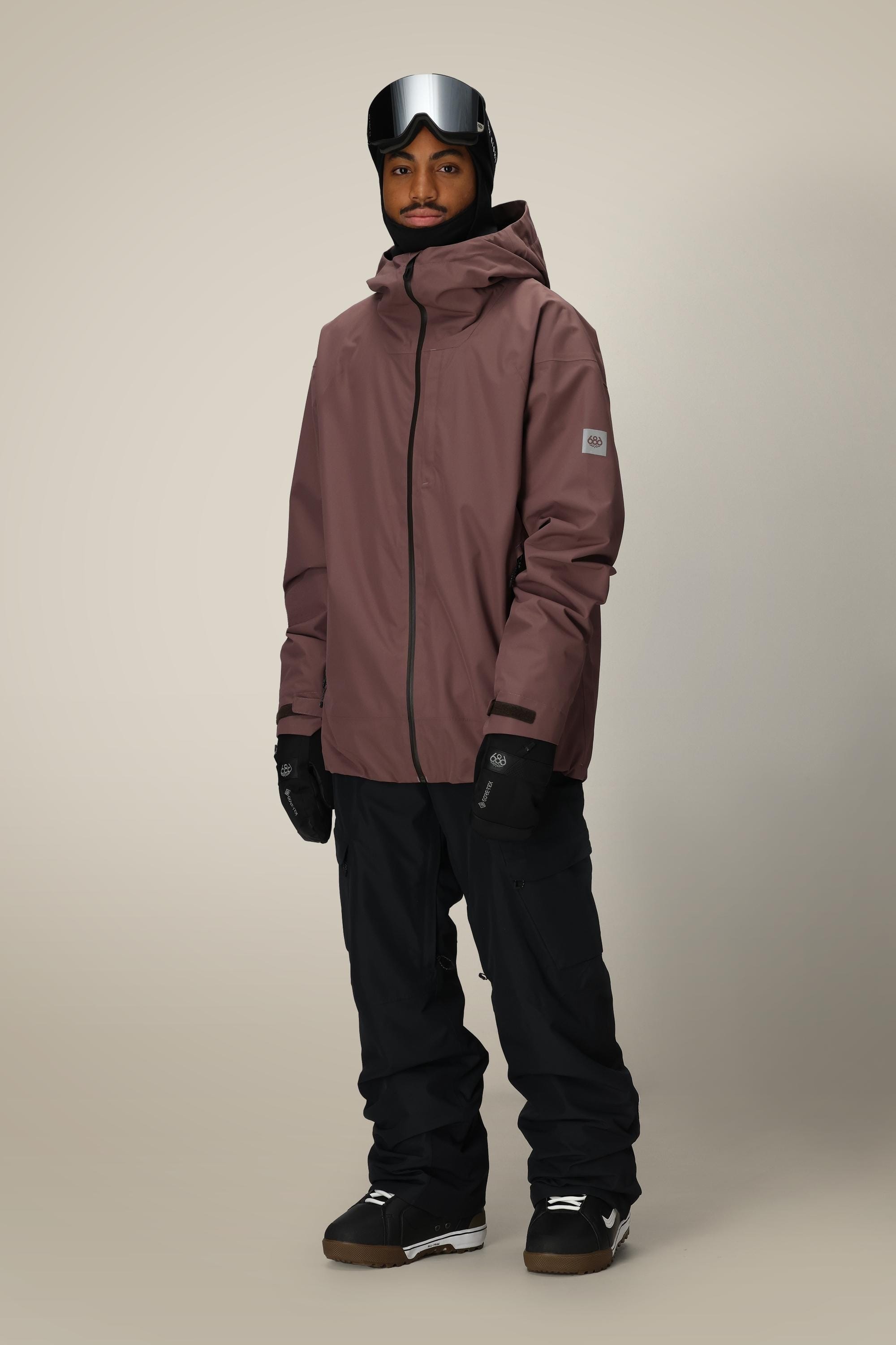 A person wearing a 686 Mens Gateway Shell Jacket in brown with a hood, black snow pants, gloves, boots, and ski goggles stands against a plain beige background.