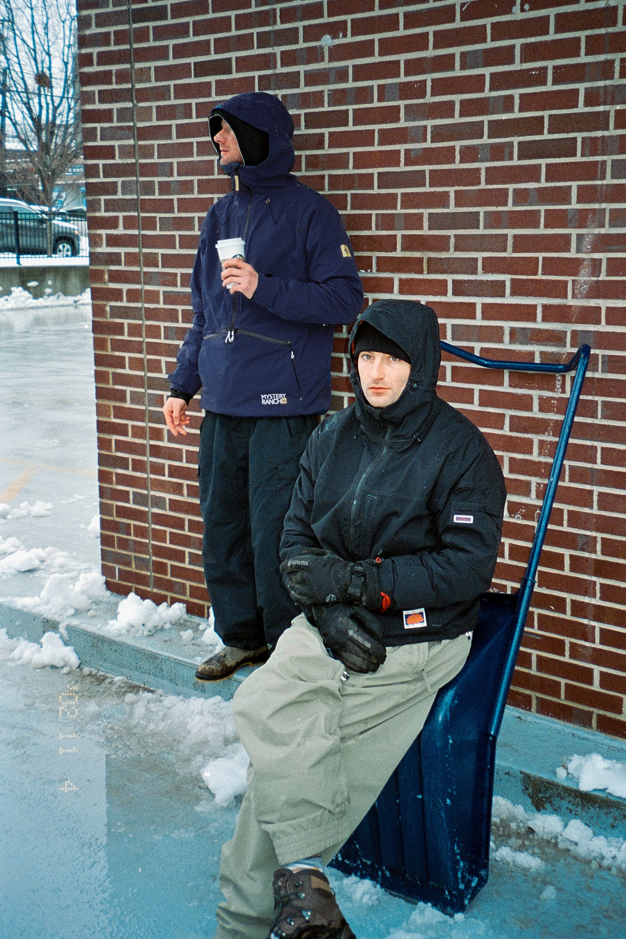 Two people in winter clothes are outside by a brick wall—one is standing in the 686 Men's Mystery Ranch Qi 3L Shell Jacket holding a coffee cup, while the other sits on a snow shovel. Snow and ice cover the ground around them.