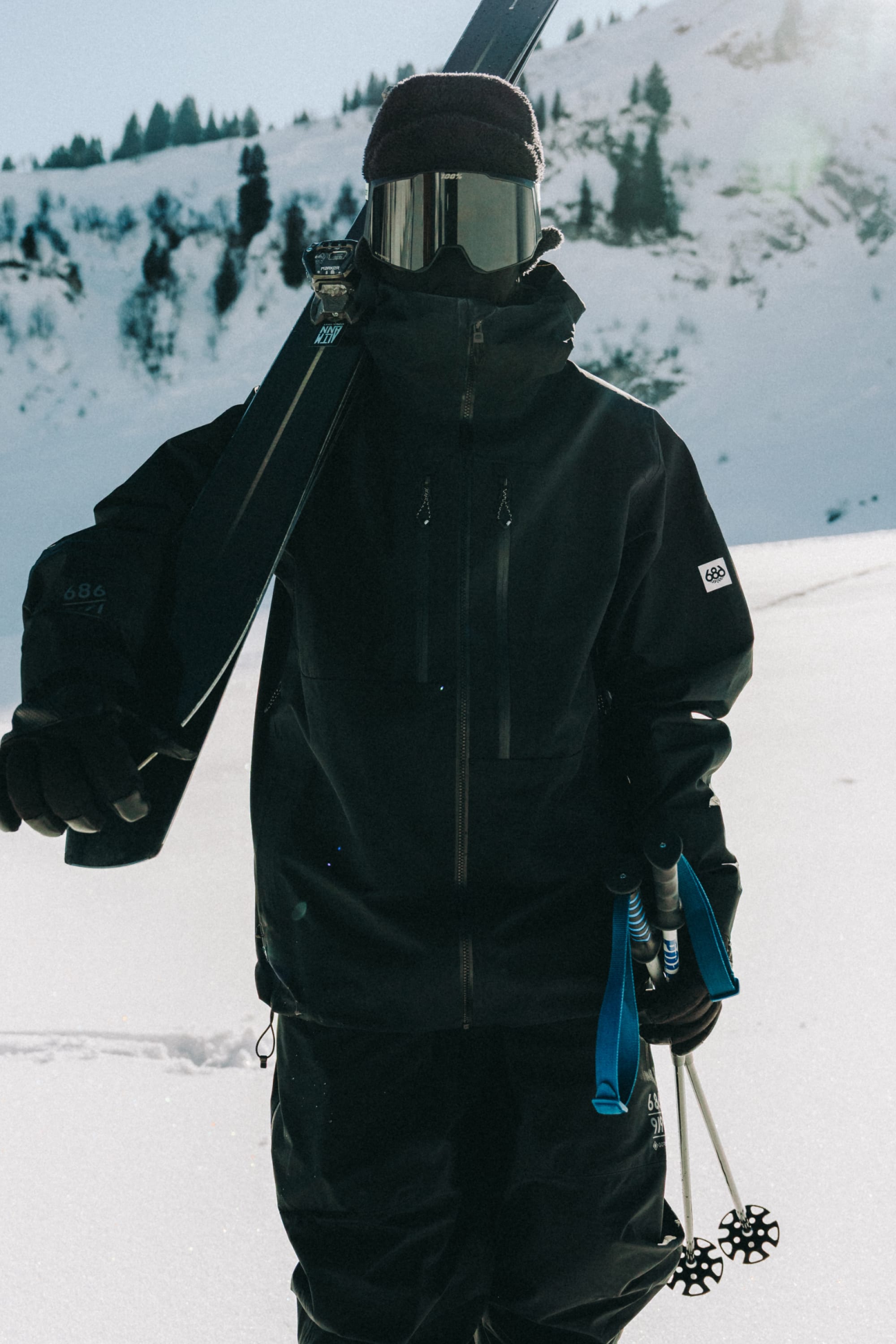 A skier wearing the 686 Men's GORE-TEX 3L ATV Thermagraph® Jacket stands on snowy slopes, holding skis and poles, with snow-covered mountains in the background.