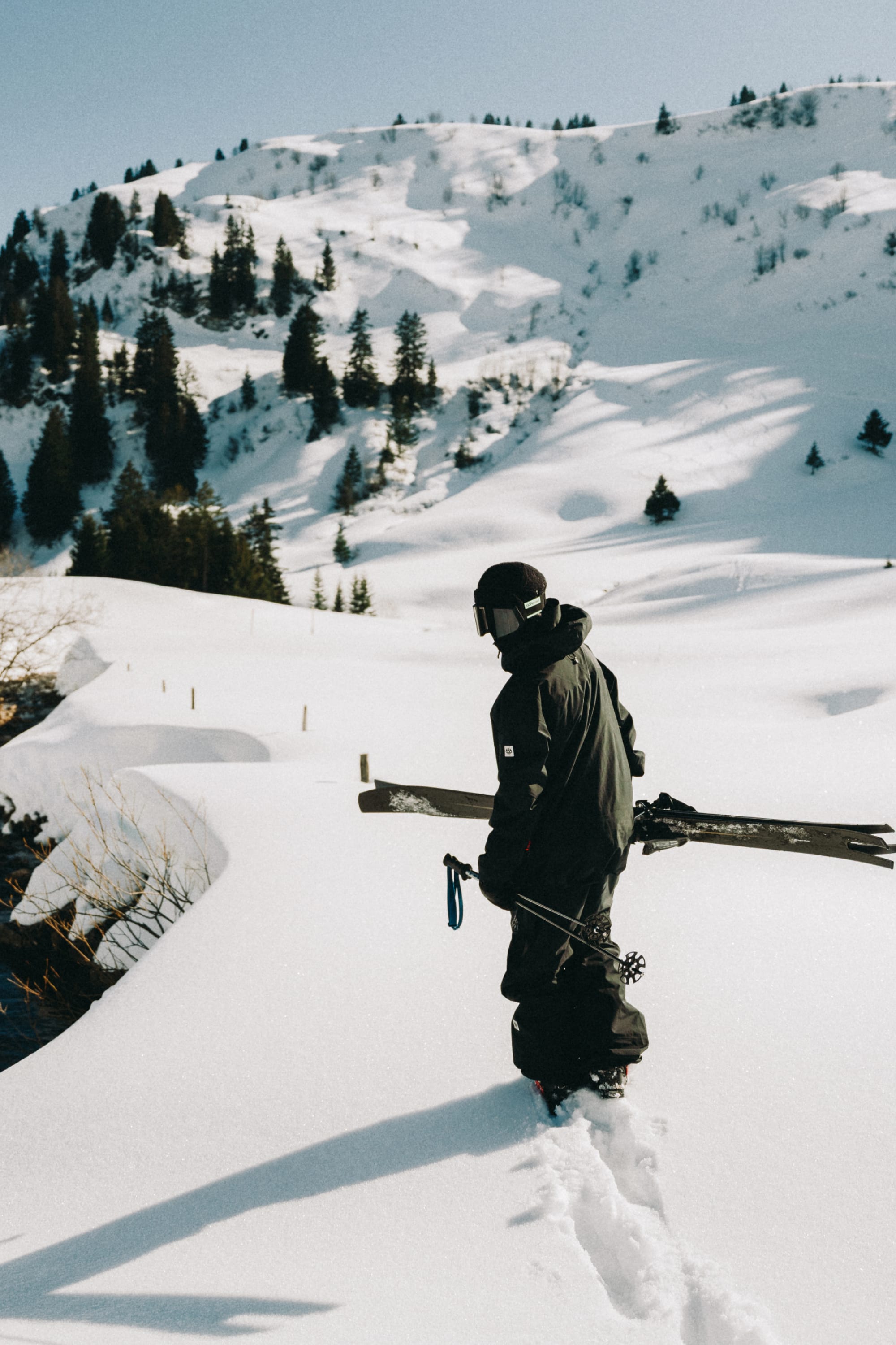 Wearing the 686 Men's GORE-TEX 3L ATV Thermagraph® Jacket, a person with a helmet carries skis while trekking through deep snow in a snowy mountain landscape dotted with pine trees.