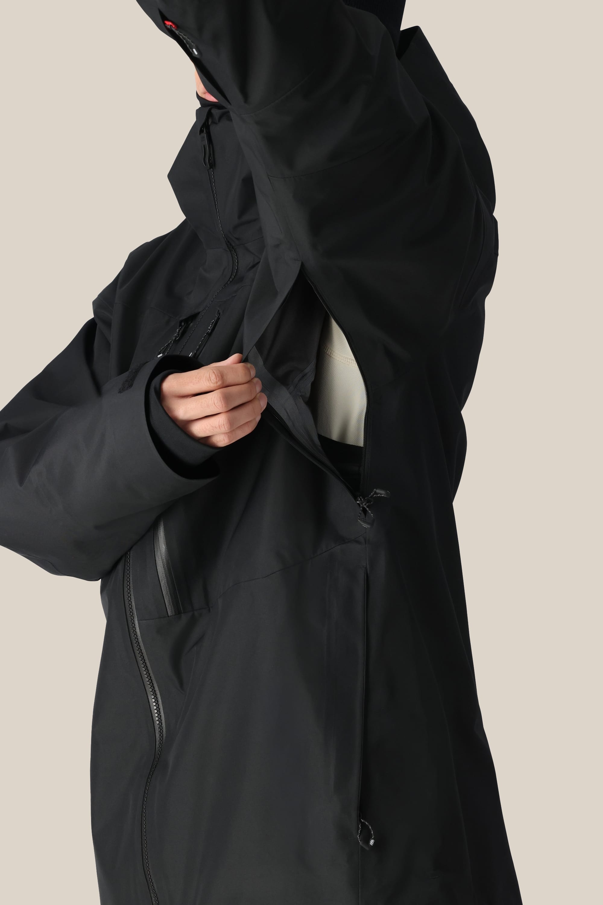 A man in a 686 Men's GORE-TEX 3L ATV Thermagraph® Jacket unzips an underarm vent, showing part of a light inner layer. The black polyester jacket by 686 features multiple zippers and a high collar.