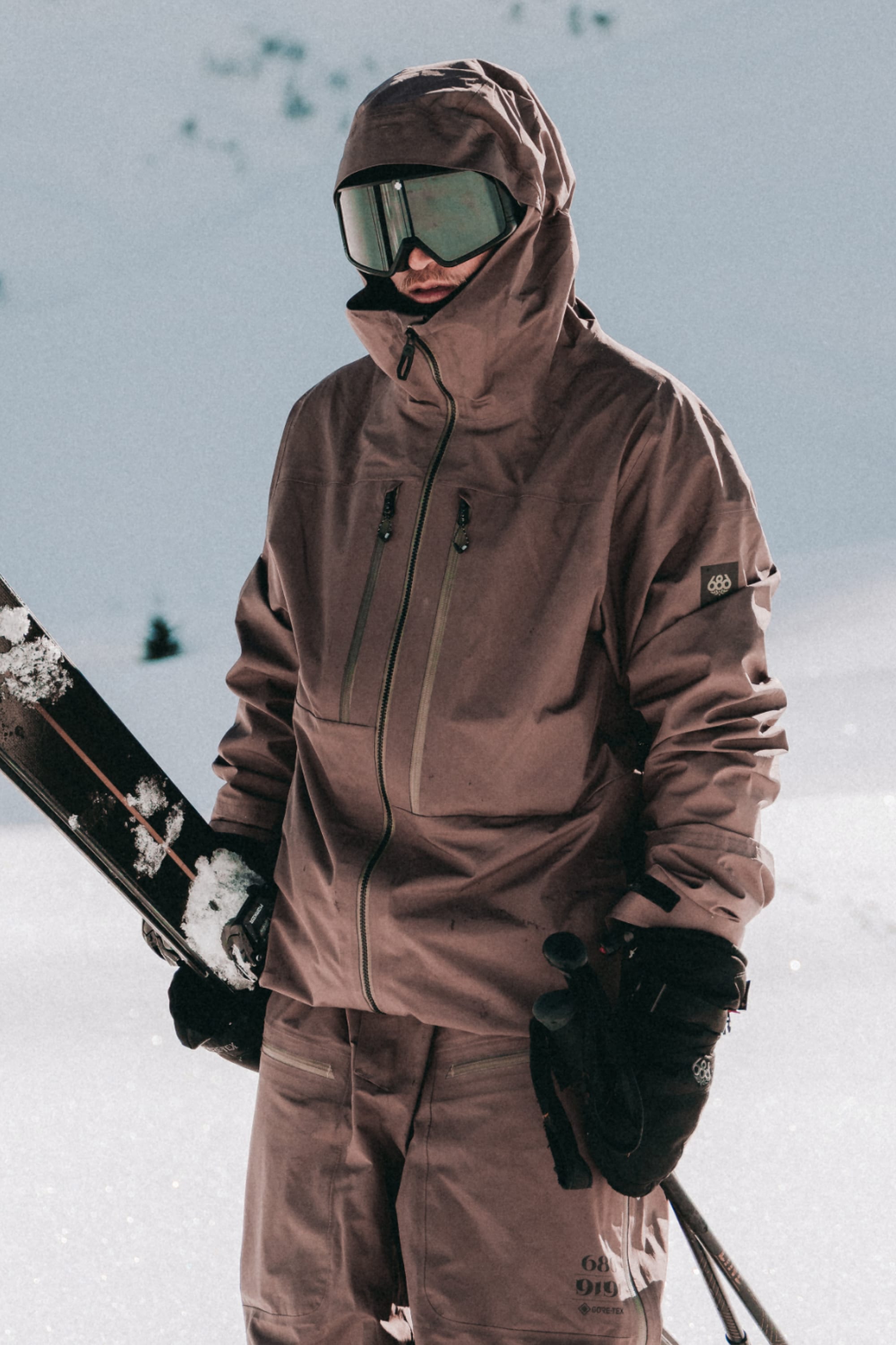 A person wearing the 686 Men's GORE-TEX 3L ATV Thermagraph® Jacket stands in the snow with skis and poles, dressed for skiing with pants, gloves, and reflective goggles. The snow-covered ground and blurred mountains are in the background.