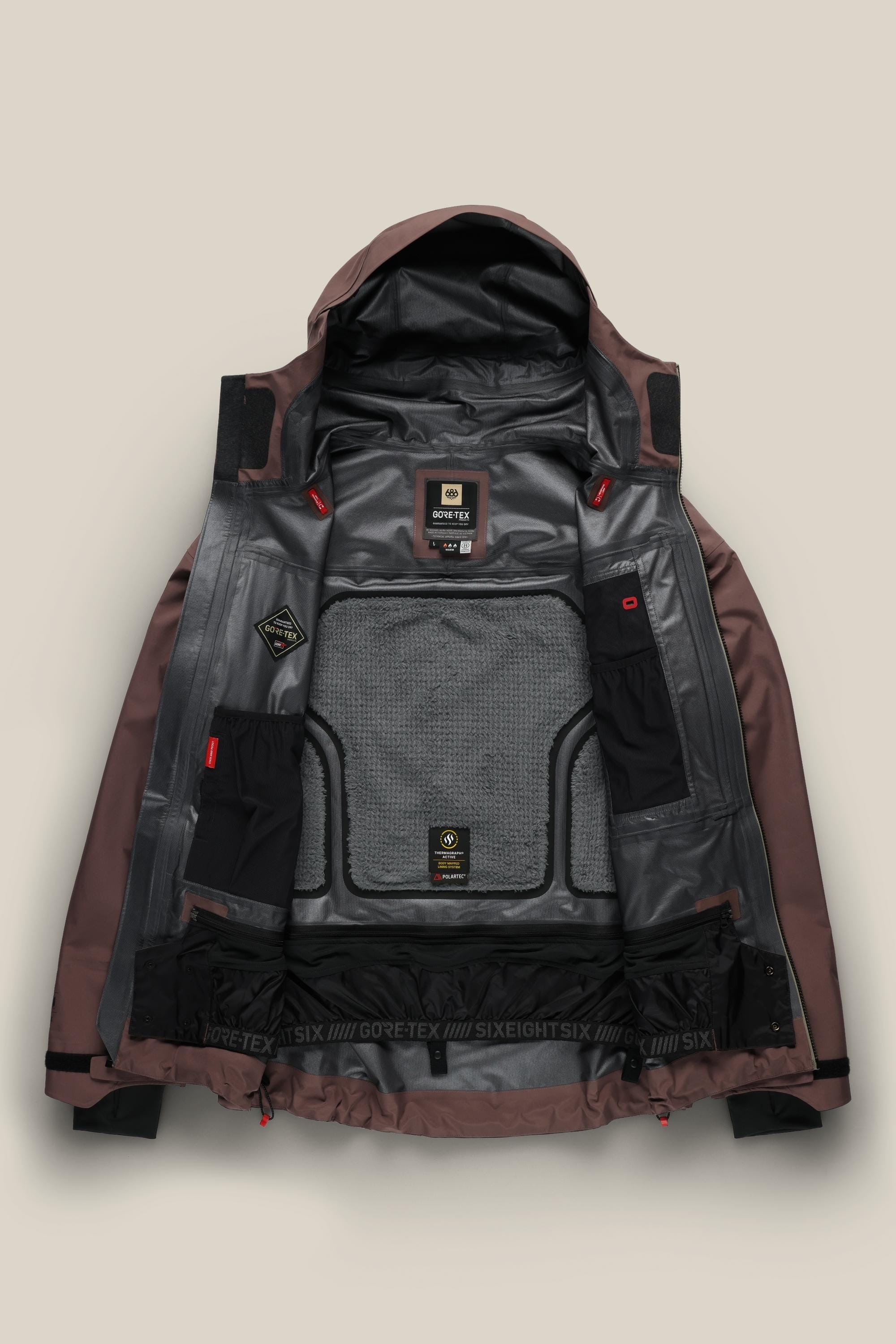 The 686 Men's GORE-TEX 3L ATV Thermagraph® Jacket in maroon and gray is laid open on a beige background, showing its black and gray insulated panels, mesh pockets, and multiple Gore-Tex and brand label tags.