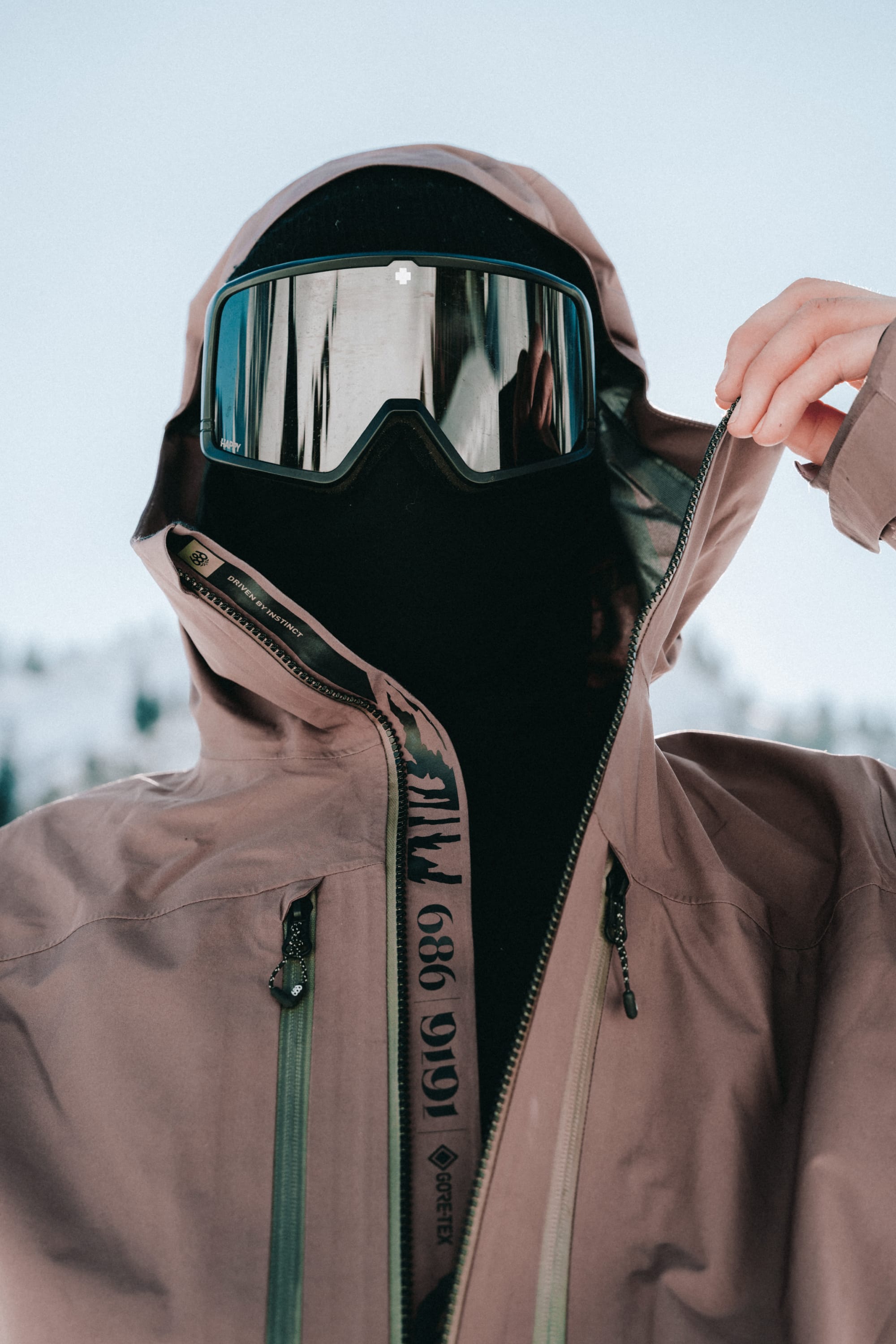 A person outdoors in snowy mountains partially unzips their brown 686 Men's GORE-TEX 3L ATV Thermagraph® Jacket, wearing a black ski mask and reflective ski goggles.