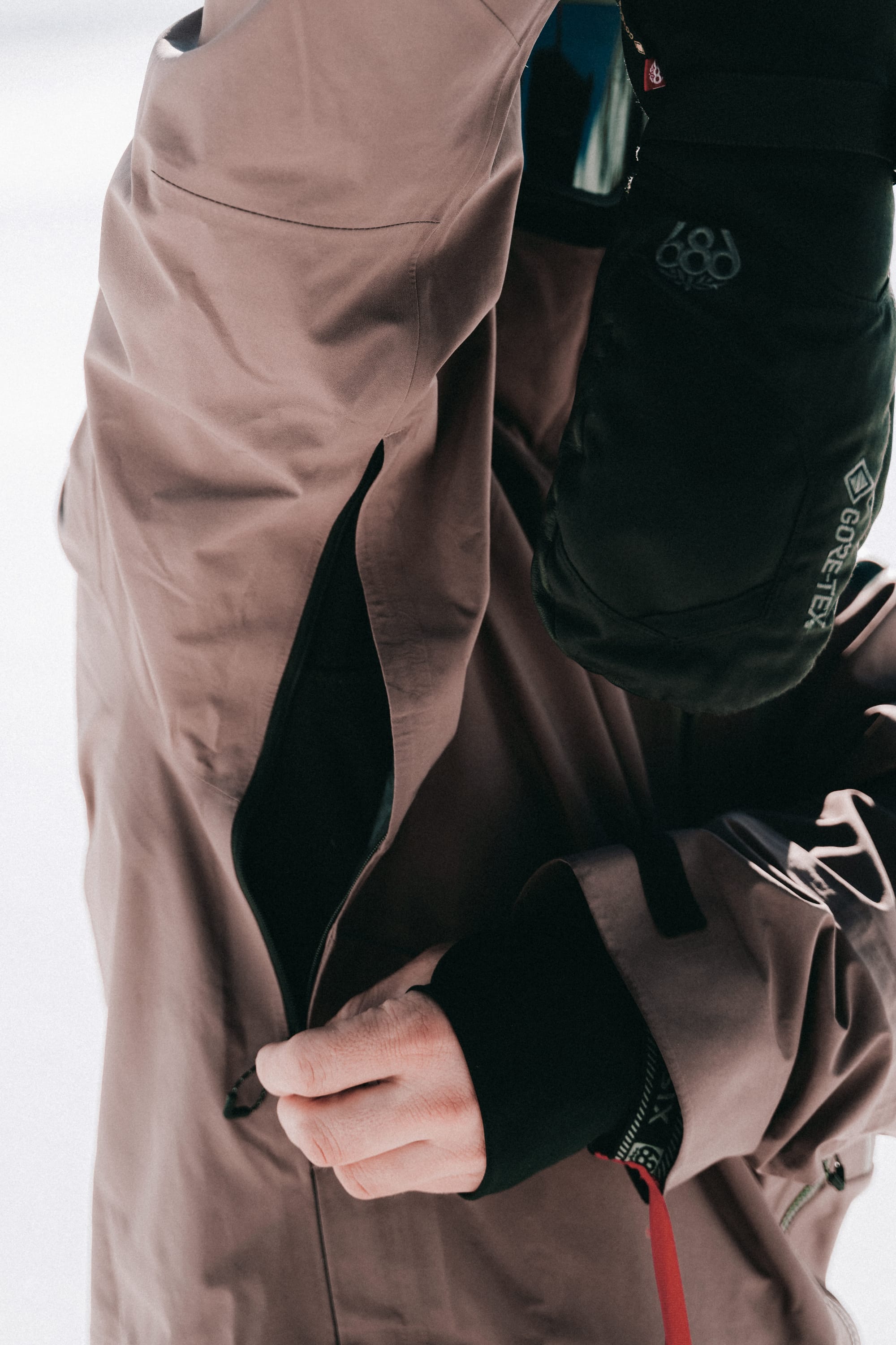 A person wearing the 686 Men's GORE-TEX 3L ATV Thermagraph® Jacket and black gloves is unzipping a sleeve pocket, with their hand and the zipper in focus while the background remains blurred.