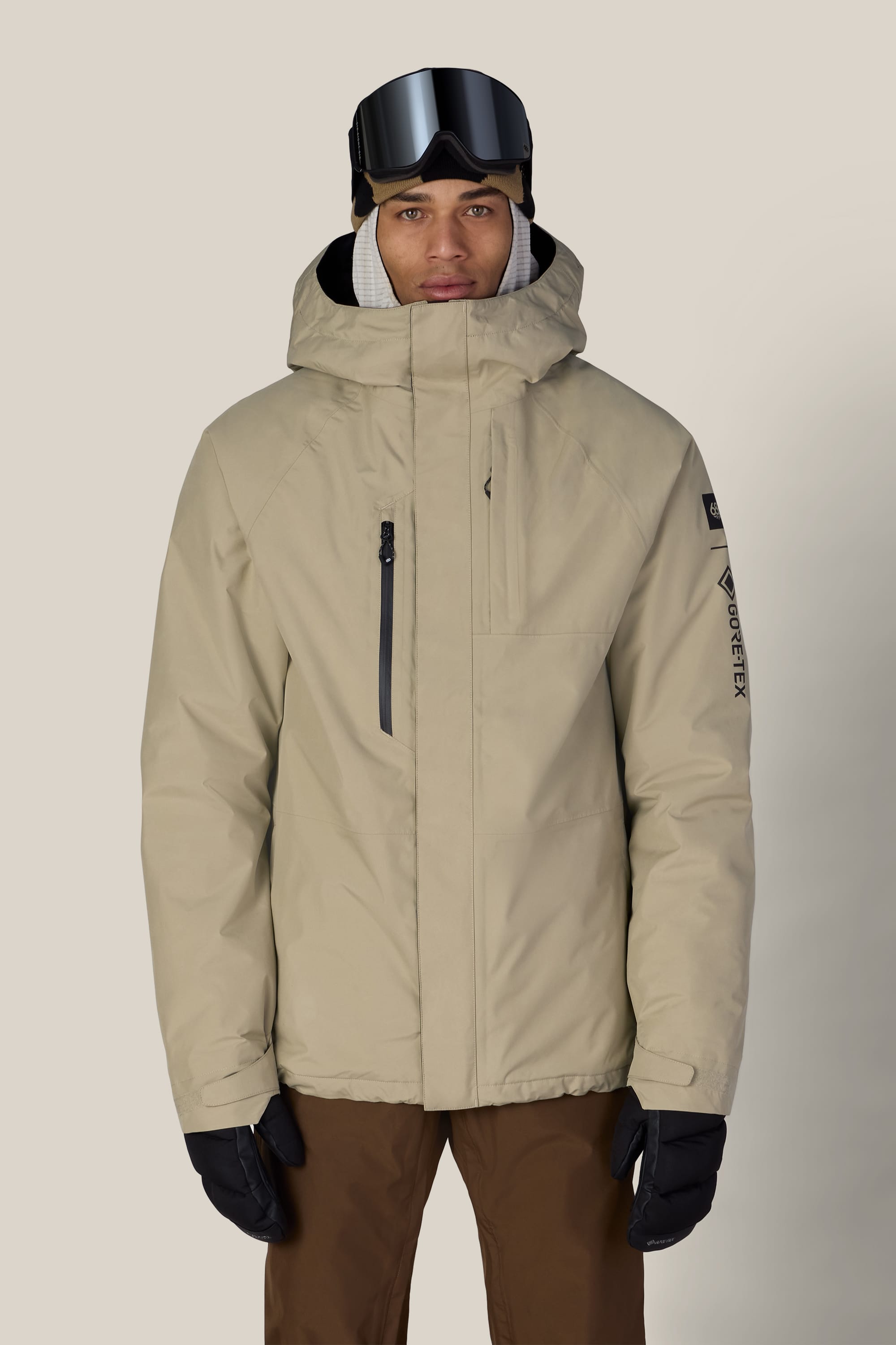 A person wearing the 686 Men’s GORE-TEX® Core Insulated Jacket by 686, paired with brown pants, black gloves, and ski goggles, stands against a plain, light background.