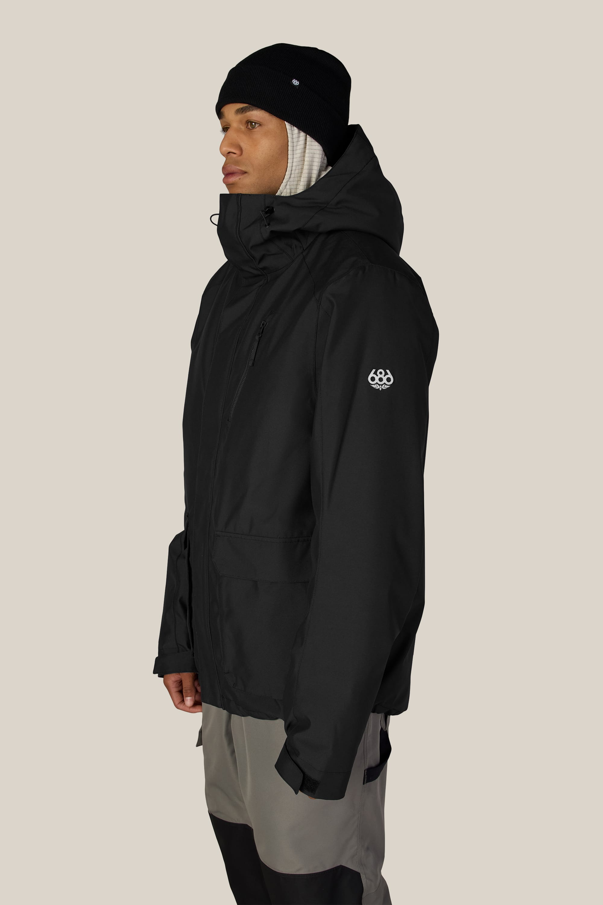 Wearing the 686 Men’s Nightline™ Insulated Jacket in black, along with a matching black beanie and beige scarf, a person stands in profile against a plain light background.