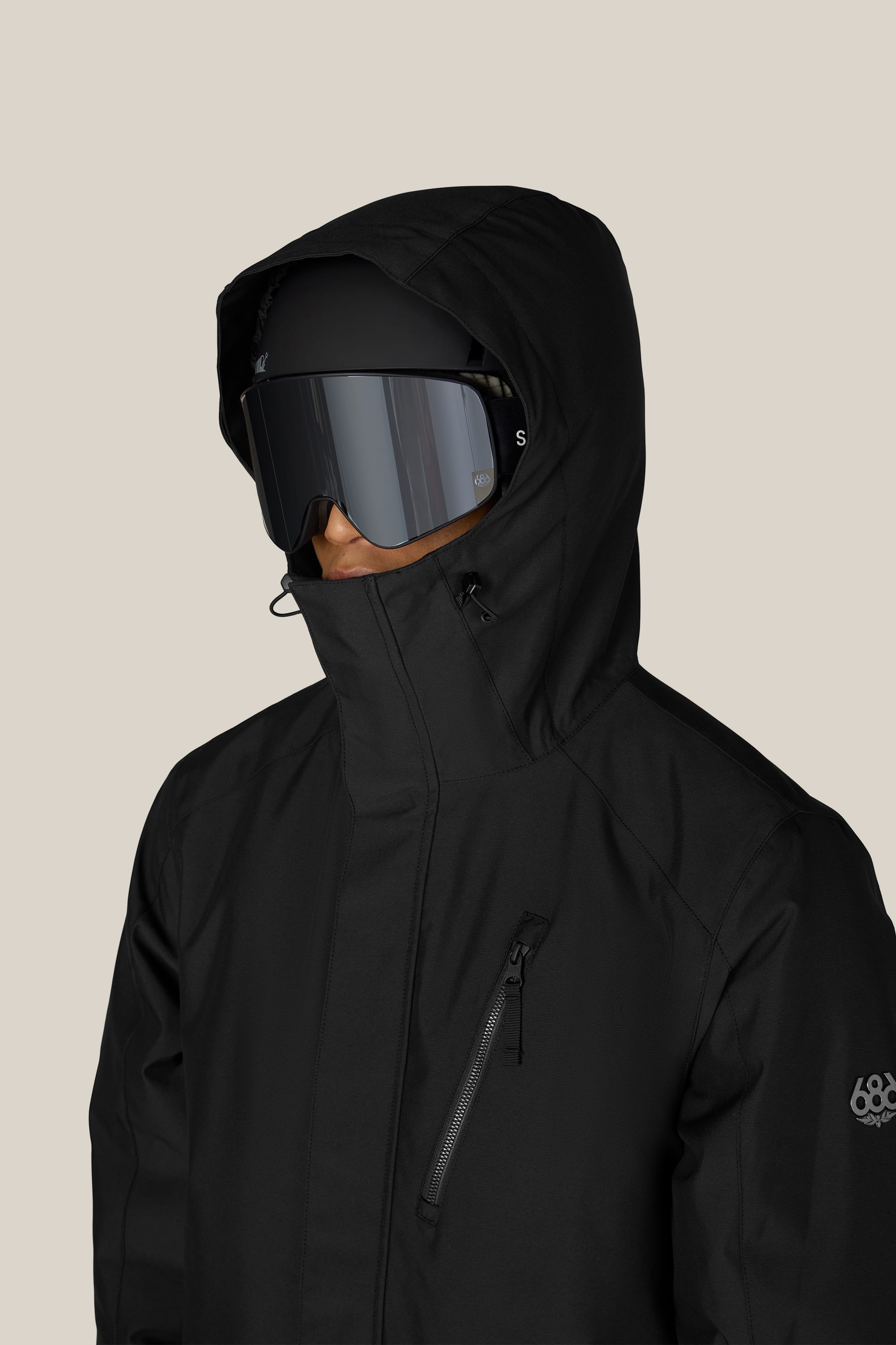 A person wearing the 686 Men’s Nightline™ Insulated Jacket, paired with large black ski goggles and a helmet, standing against a plain light background.