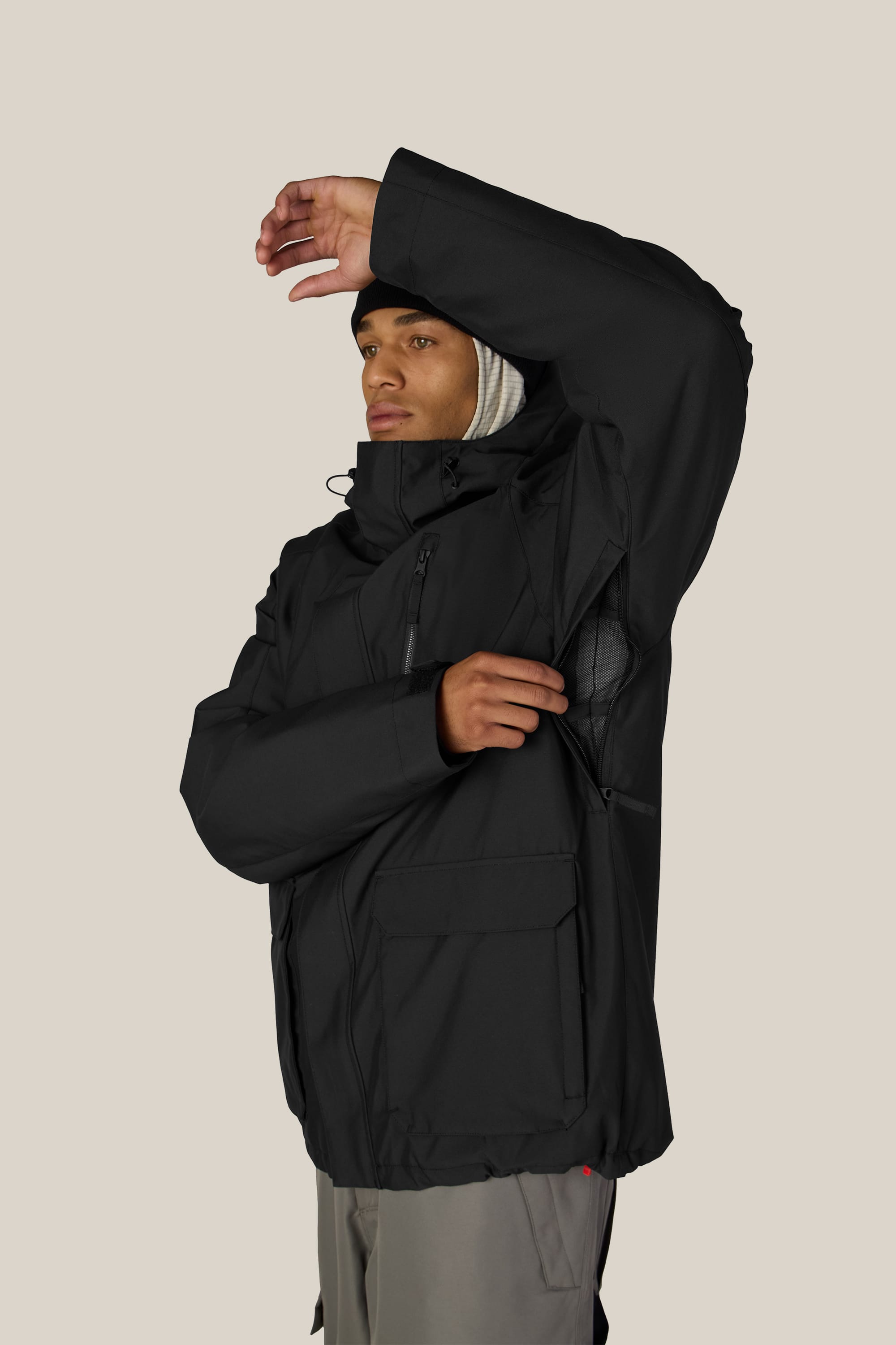 A person wearing the 686 Men’s Nightline™ Insulated Jacket by 686 raises one arm to unzip the jacket’s side vent, exposing its interior lining against a plain, light-colored background.