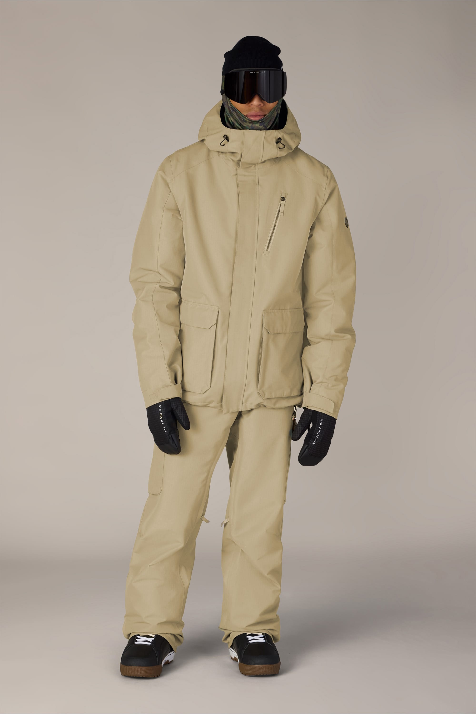 A person wearing the 686 Men’s Nightline™ Insulated Jacket by 686, paired with matching tan pants, black gloves, black boots, a black beanie, dark ski goggles, and a green neck gaiter, standing against a light neutral background.