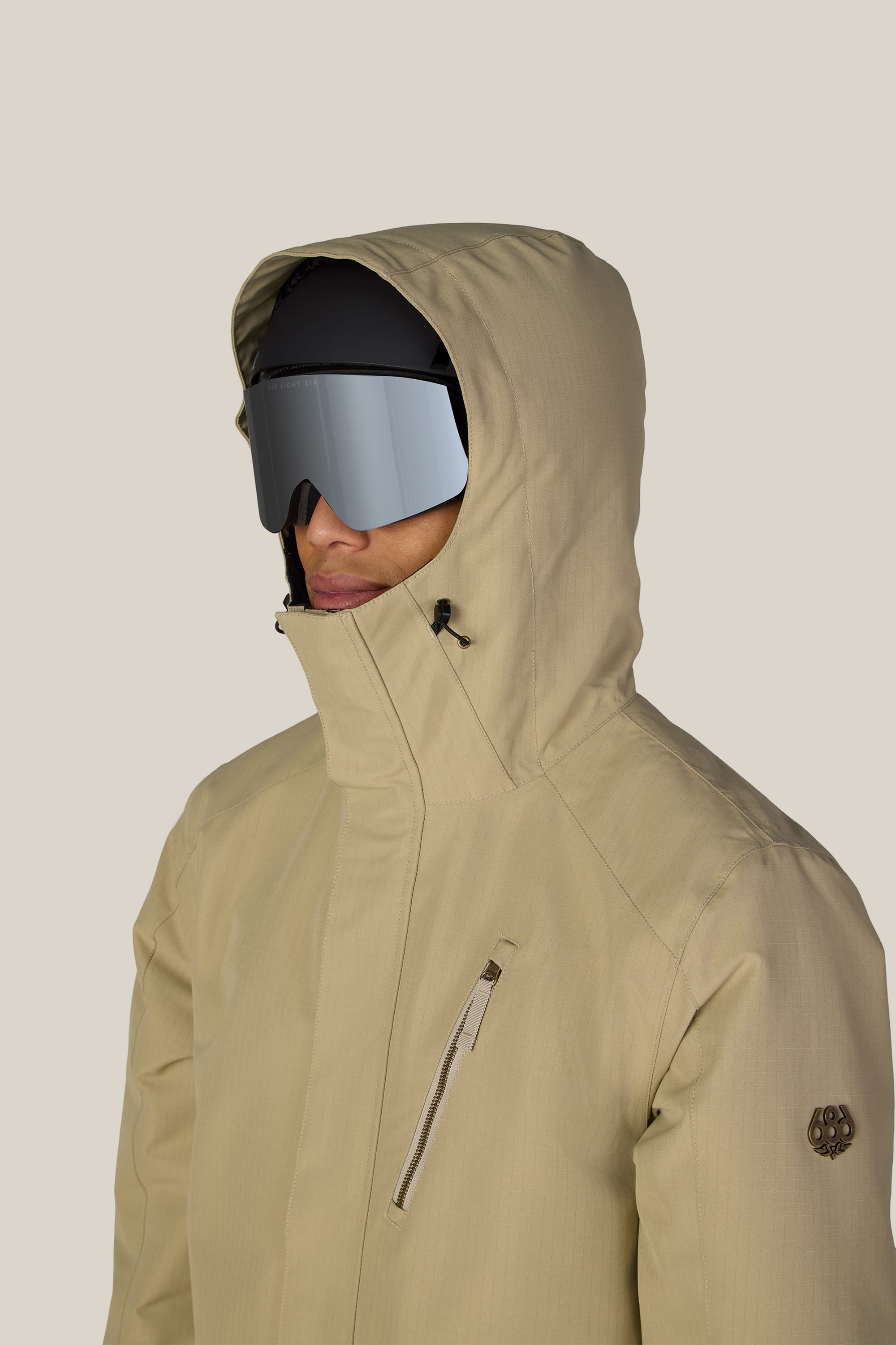 A person is wearing the 686 Men’s Nightline™ Insulated Jacket in beige, paired with black ski goggles and a black cap, facing slightly left. The jacket by 686 features a zippered chest pocket and an emblem on the left sleeve. The background is plain light gray.