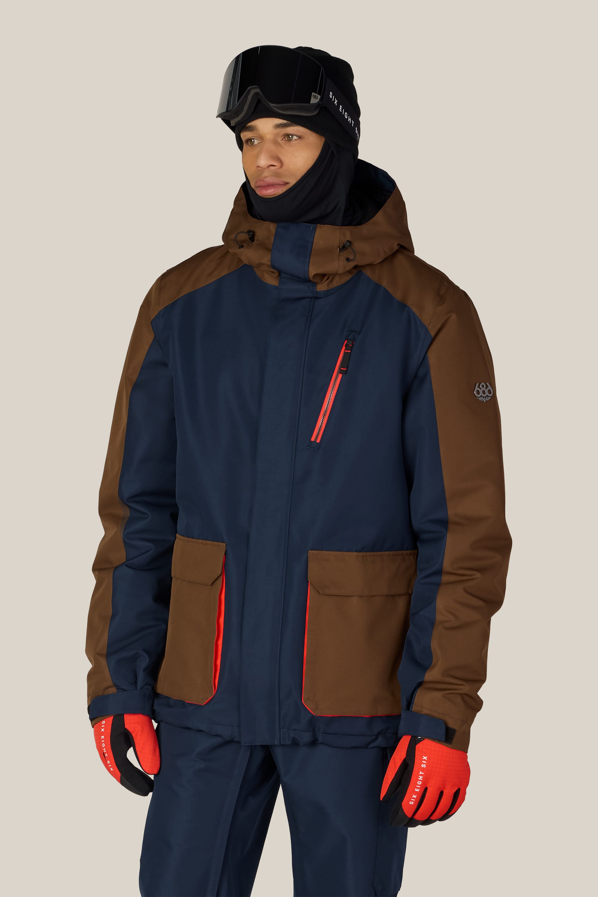 A person wearing the 686 Men’s Nightline™ Insulated Jacket by 686 in navy and brown, along with red gloves, navy pants, ski goggles, and a black balaclava stands against a plain light background.