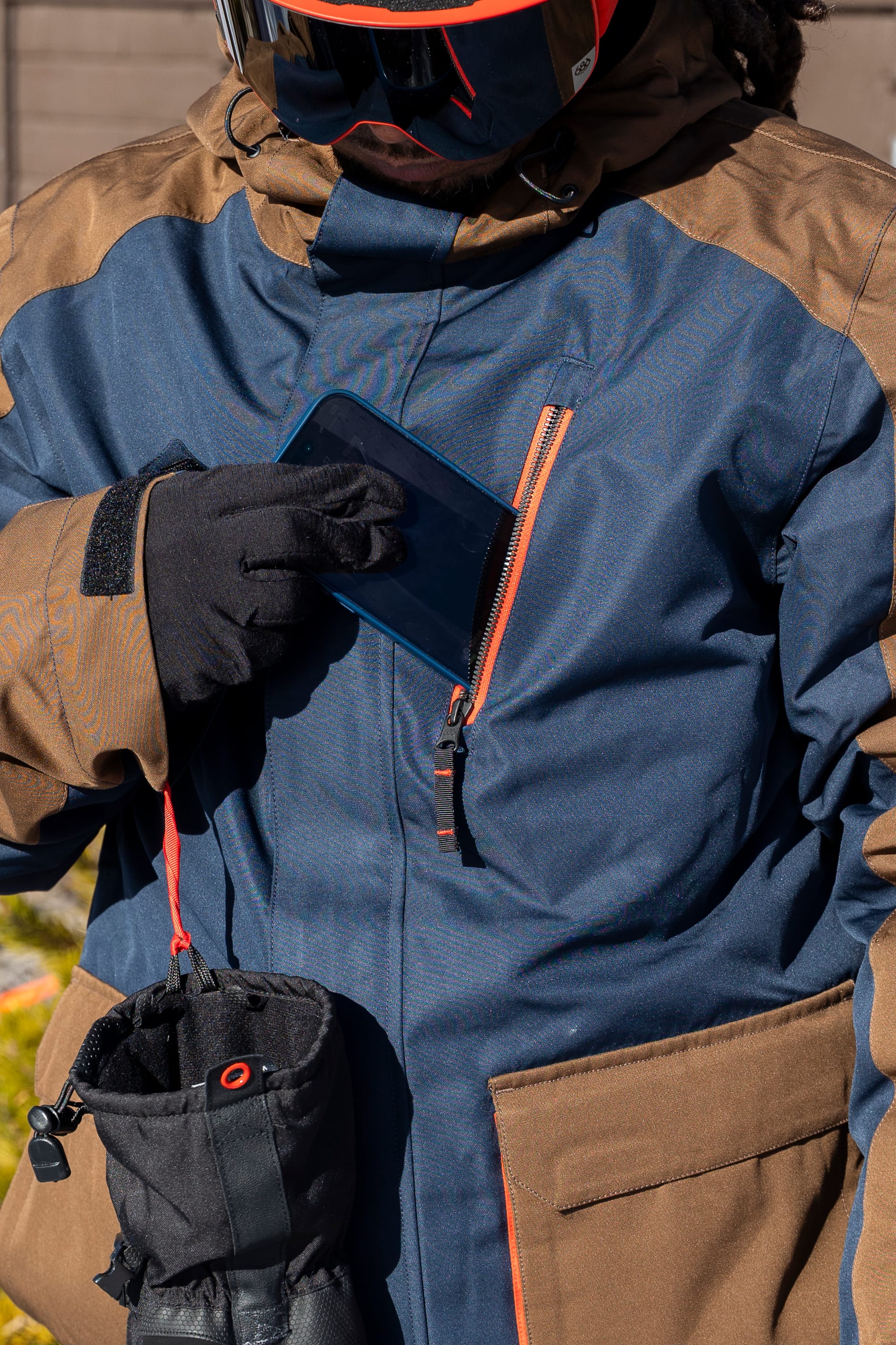 A person wearing the 686 Men’s Nightline™ Insulated Jacket in blue and brown, along with black gloves, puts a phone into their chest pocket. A ski pass holder and part of a glove are attached to their wrist.