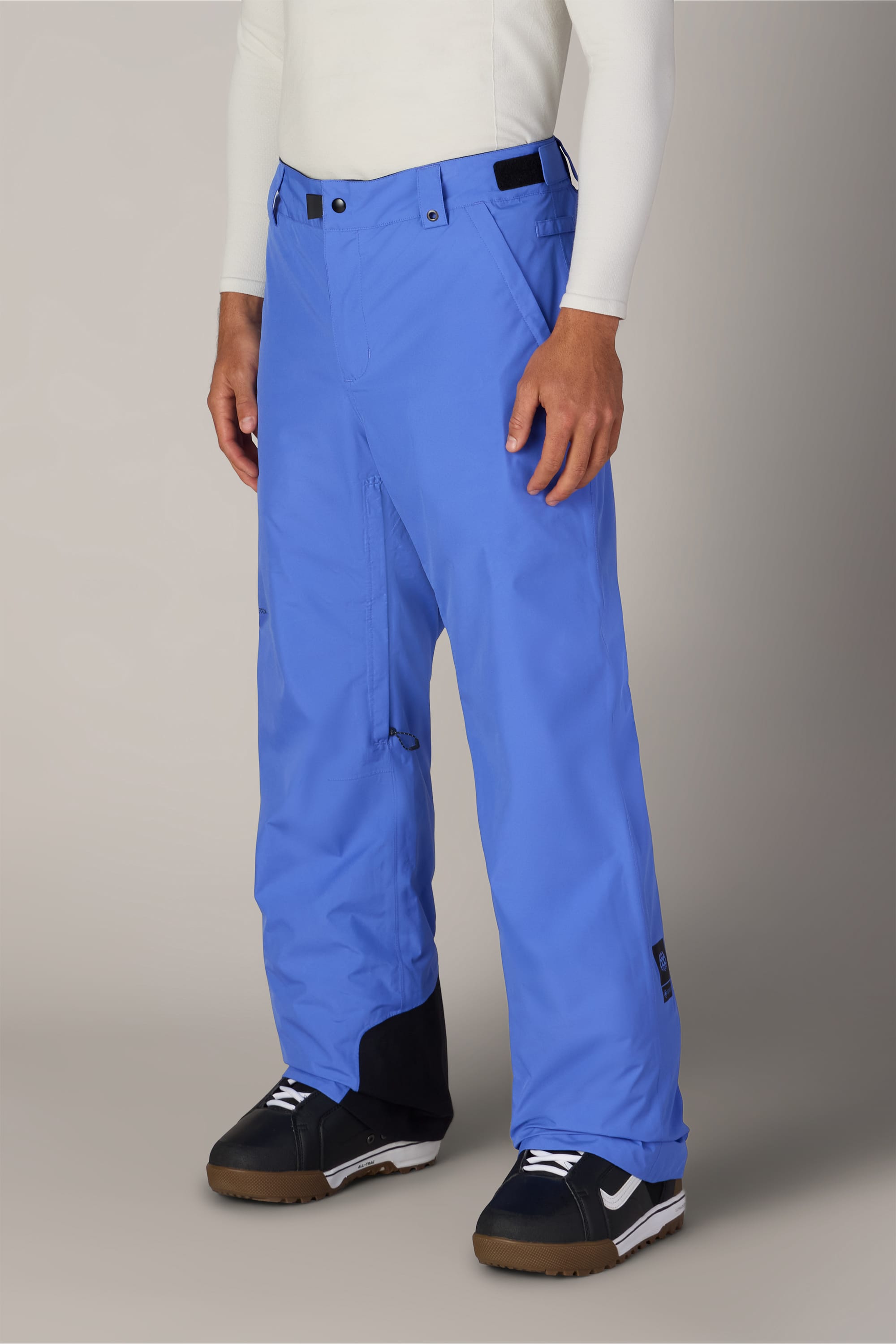 A person wearing 686 Men’s GORE-TEX® Core Shell Pants in bright blue, paired with a white long-sleeve top and black-and-white sneakers, stands against a plain light background. Only the lower body is visible.