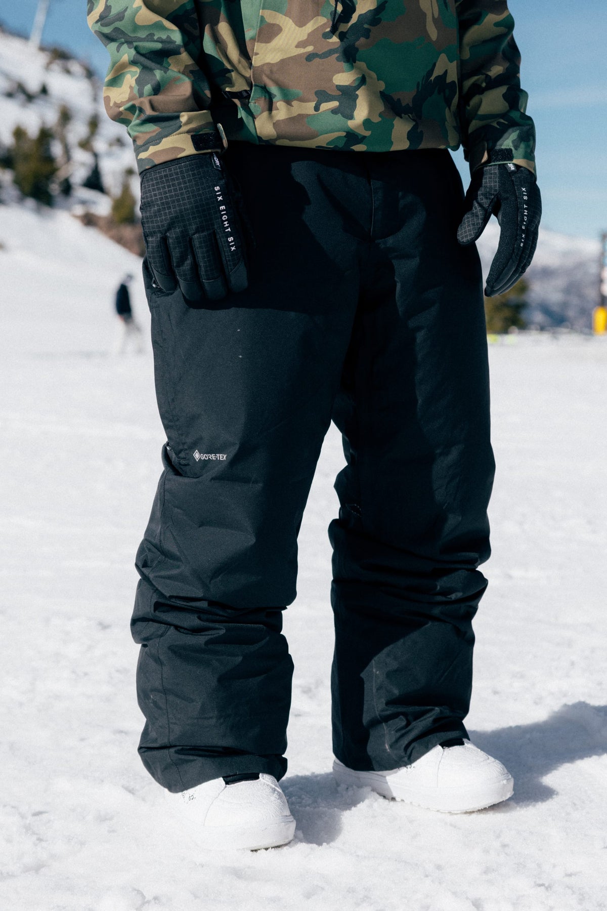 A person wearing 686 Men’s GORE-TEX® Core Shell Pant stands on snowy ground with trees and mountains in the background.