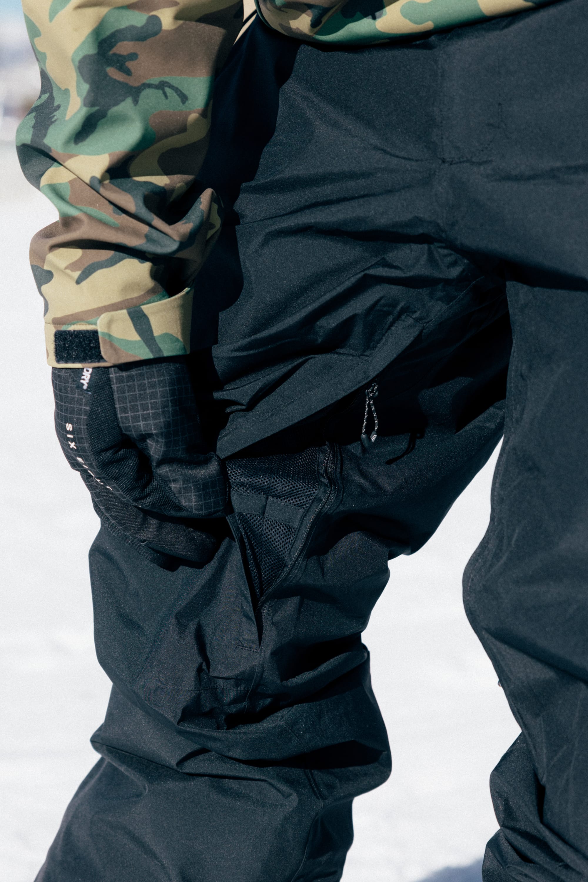 A person wearing the 686 Men’s GORE-TEX® Core Shell Pant stands outdoors on snow, holding the zippered pocket of the pants, paired with black gloves and a camouflage jacket.