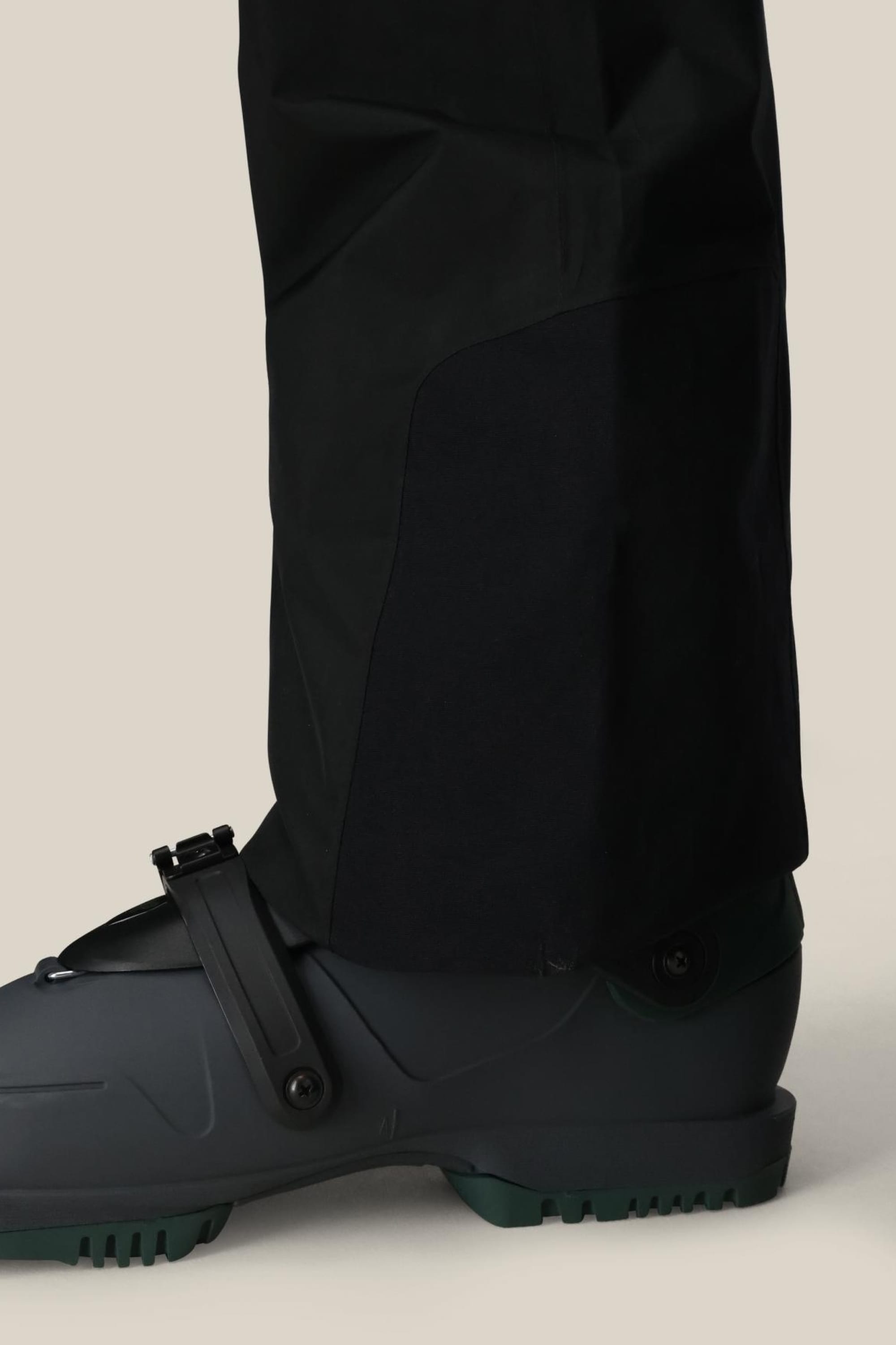 Close-up of a person’s lower leg wearing the 686 Mens GORE-TEX Core Shell Pant and a dark grey ski boot with a black strap, set against a neutral beige background.