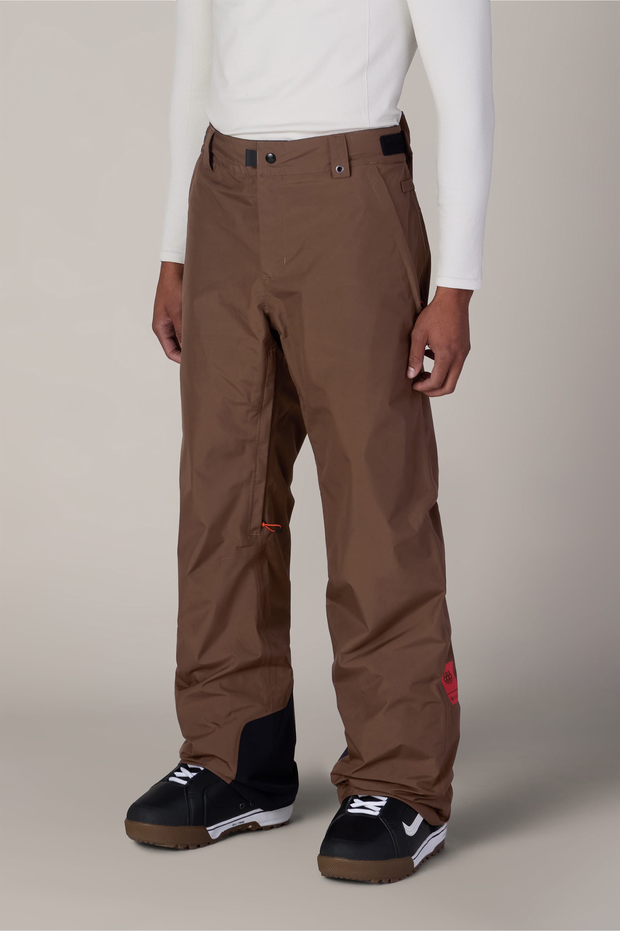 A person wearing the 686 Men’s GORE-TEX® Core Shell Pant in brown, a white long-sleeve top, and black snow boots stands against a plain background.